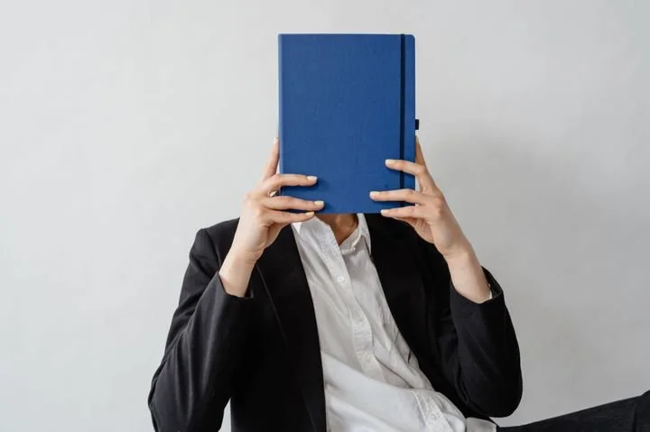 Young professional hiding behind a notebook, symbolizing identity struggles, career pressure, burnout, and self-worth issues commonly experienced by young adults navigating work and personal growth.