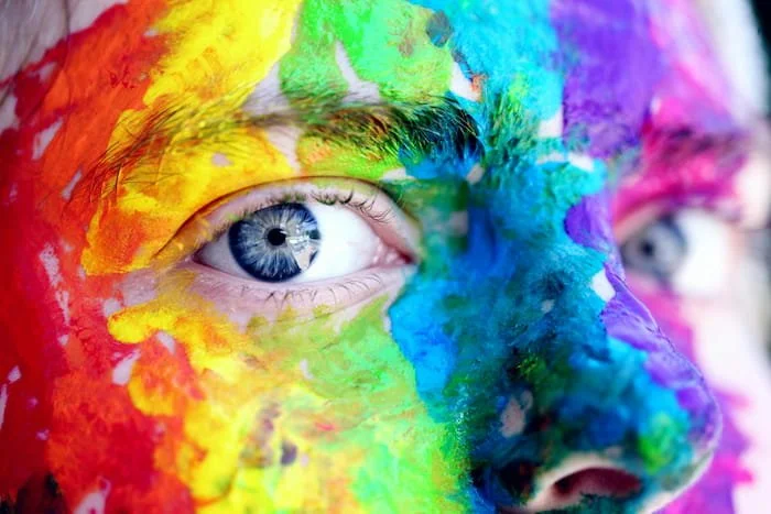 A close-up of a person's face covered in vibrant rainbow paint, with one eye clearly visible, symbolizing gender identity exploration, authenticity, and the diverse spectrum of self-expression.
