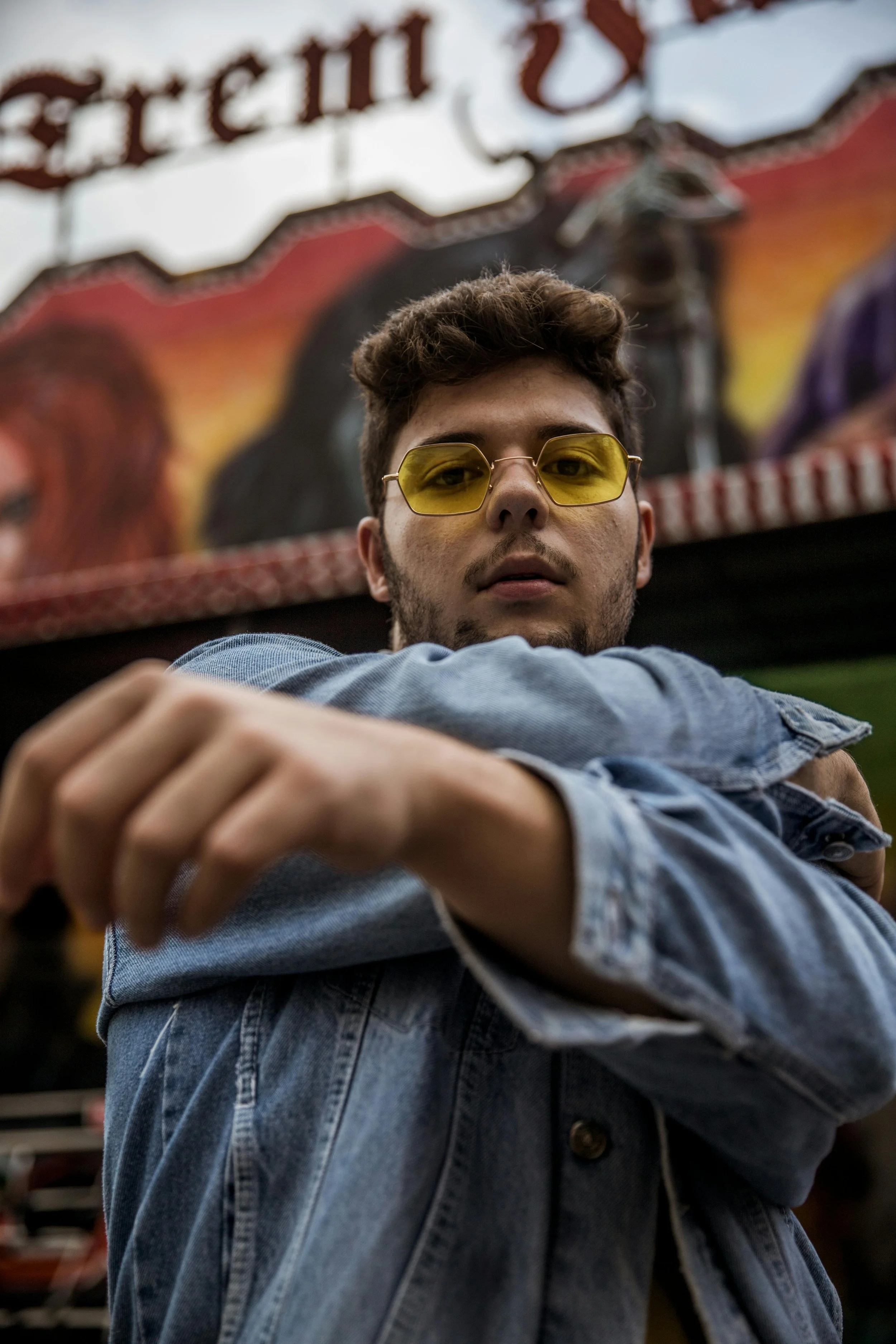 A young adult wearing yellow-tinted glasses and a denim jacket, looking directly at the camera with a guarded expression, symbolizing the emotional weight of shame and its impact on LGBTQIA+ individuals.