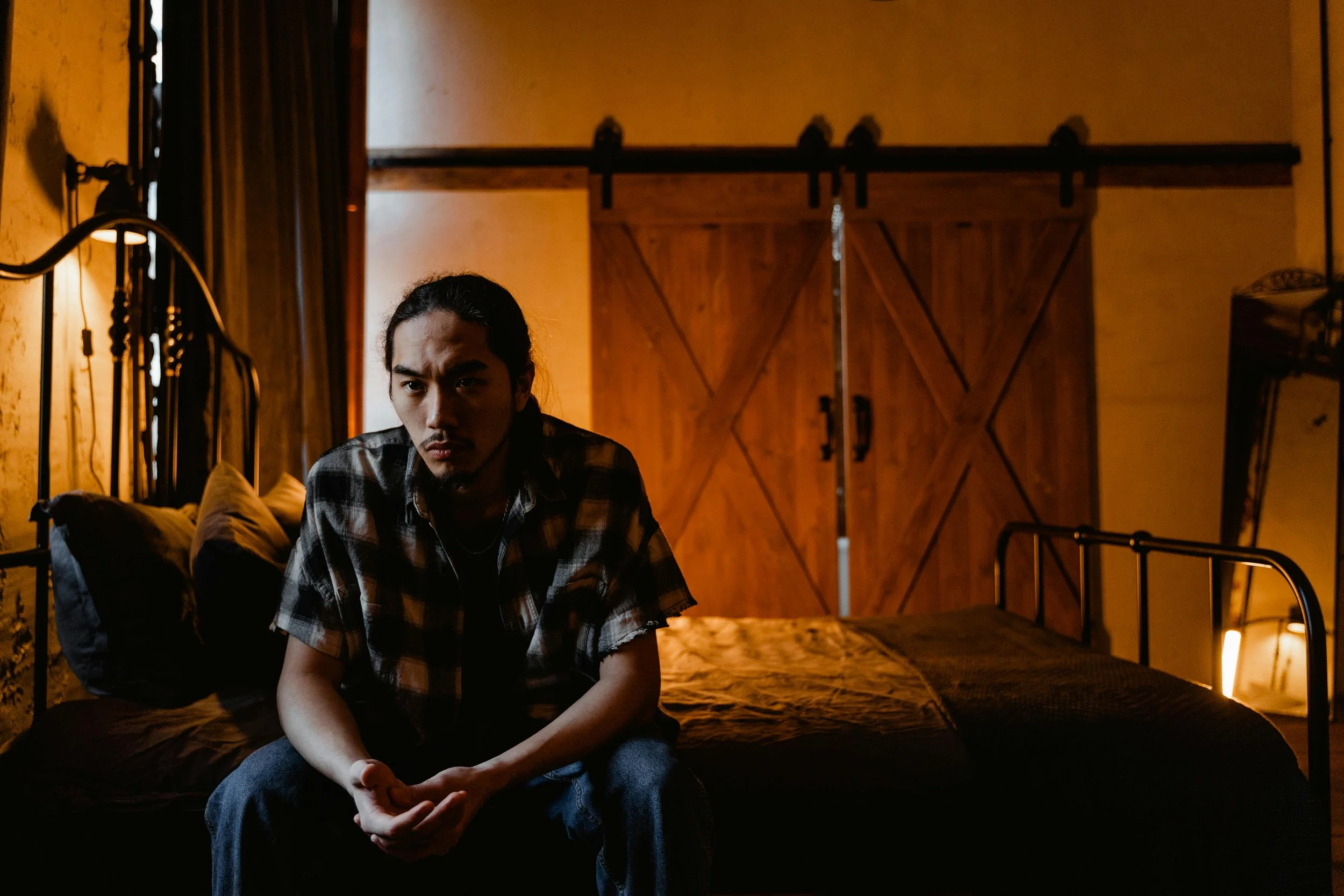 Young man sitting on the edge of a bed in a dimly lit room, looking pensive and overwhelmed.