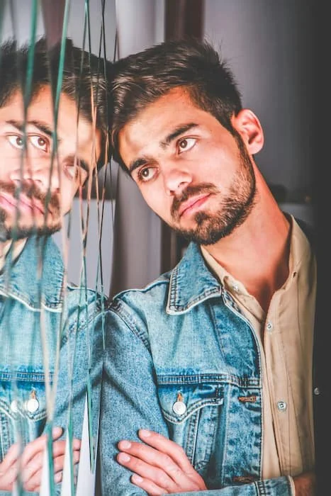 A young man leaning his head against a mirrored surface with a worried expression, symbolizing anxiety, uncertainty, and the emotional challenges of stepping into adulthood.
