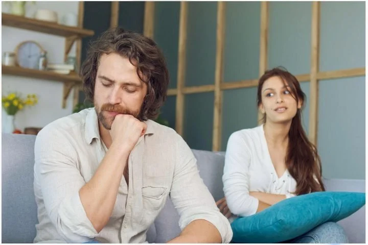 Thoughtful man looking down while his partner sits behind him on a sofa, illustrating emotional distance and sensitivity to criticism in relationships.