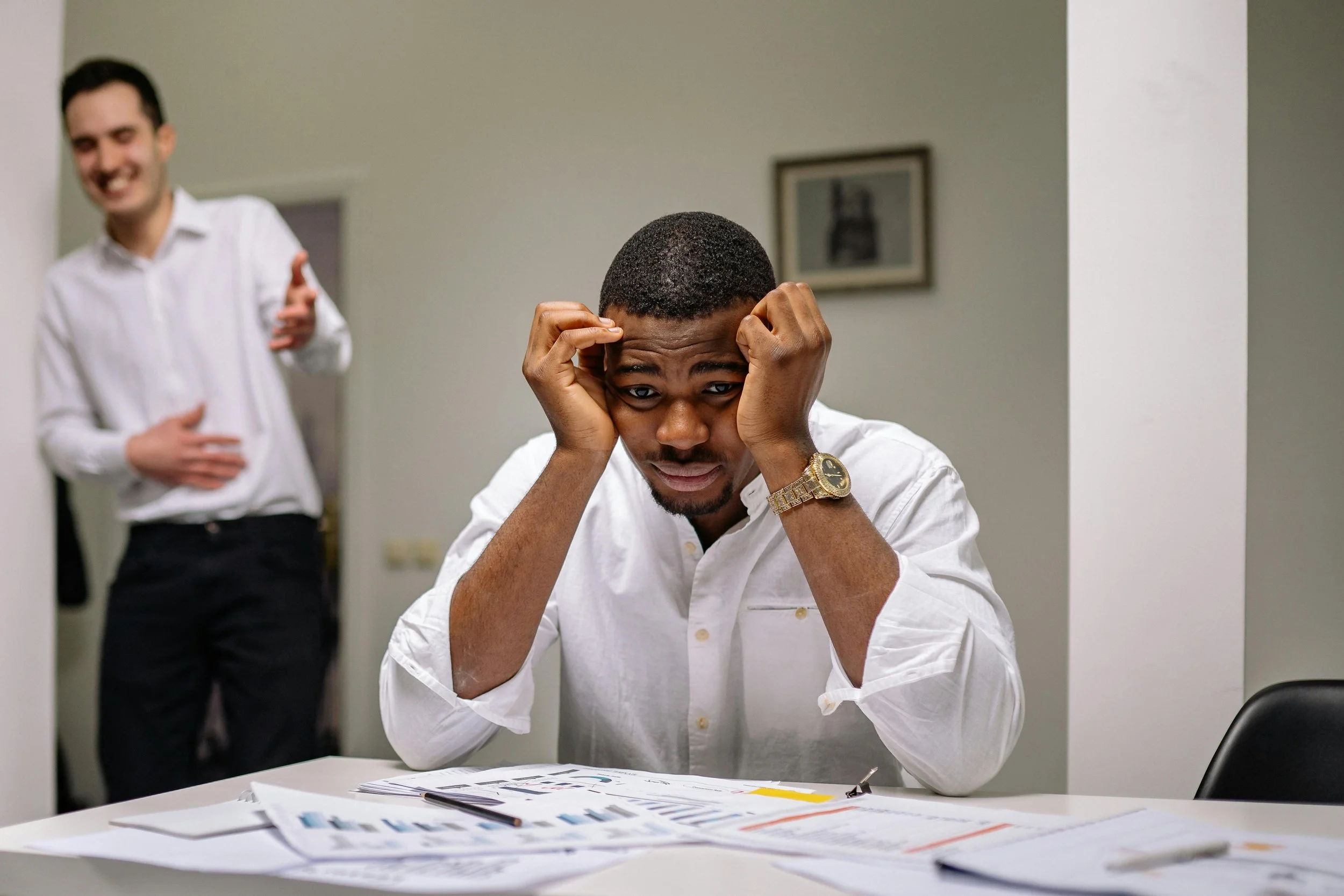 A man sitting with his head in his hands while someone speaks in the background, symbolizing the pressure and self-doubt often associated with imposter syndrome.