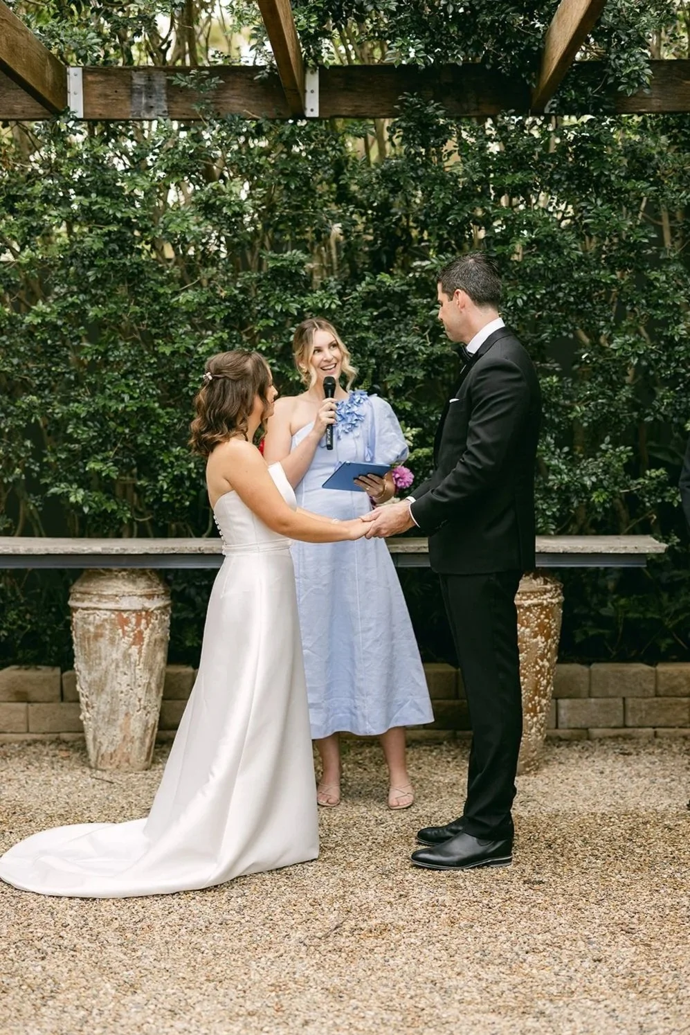 A wedding ceremony outdoors with a bride and groom holding hands and exchanging vows, Love By Britt celebrant holding a microphone, surrounded by lush greenery and decorative vases.