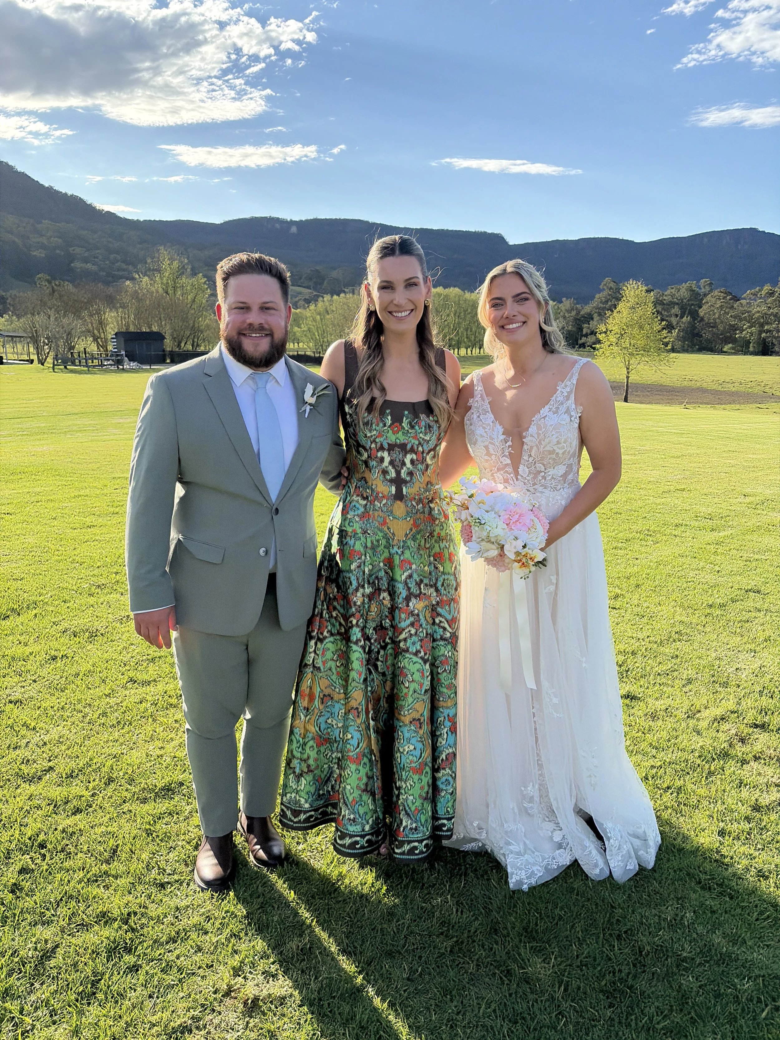 Three people standing outdoors on a grassy field with mountains and a blue sky in the background, dressed in formal attire for a wedding.