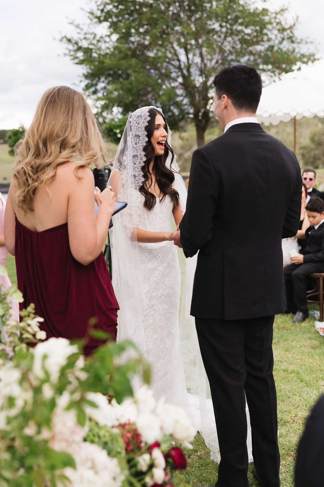 A wedding ceremony outdoors with a bride and groom holding hands, a woman officiating, and guests sitting in the background. The bride is wearing a white dress and a lace veil, the groom a black suit, and the officiant a burgundy dress, with greenery and trees surrounding them.