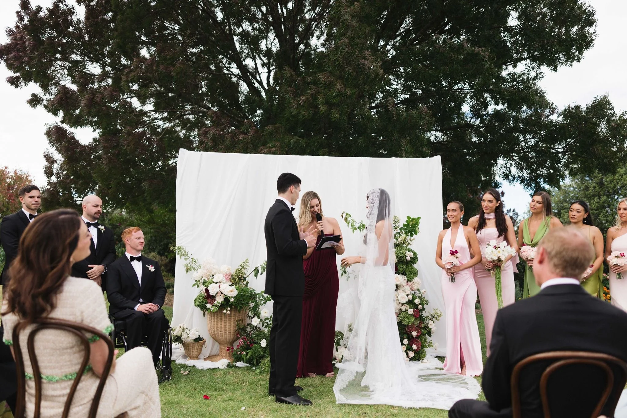 A wedding ceremony outdoors, with a bride and groom exchanging vows under a decorated arch, surrounded by friends and family. Love By Britt is the celebrant