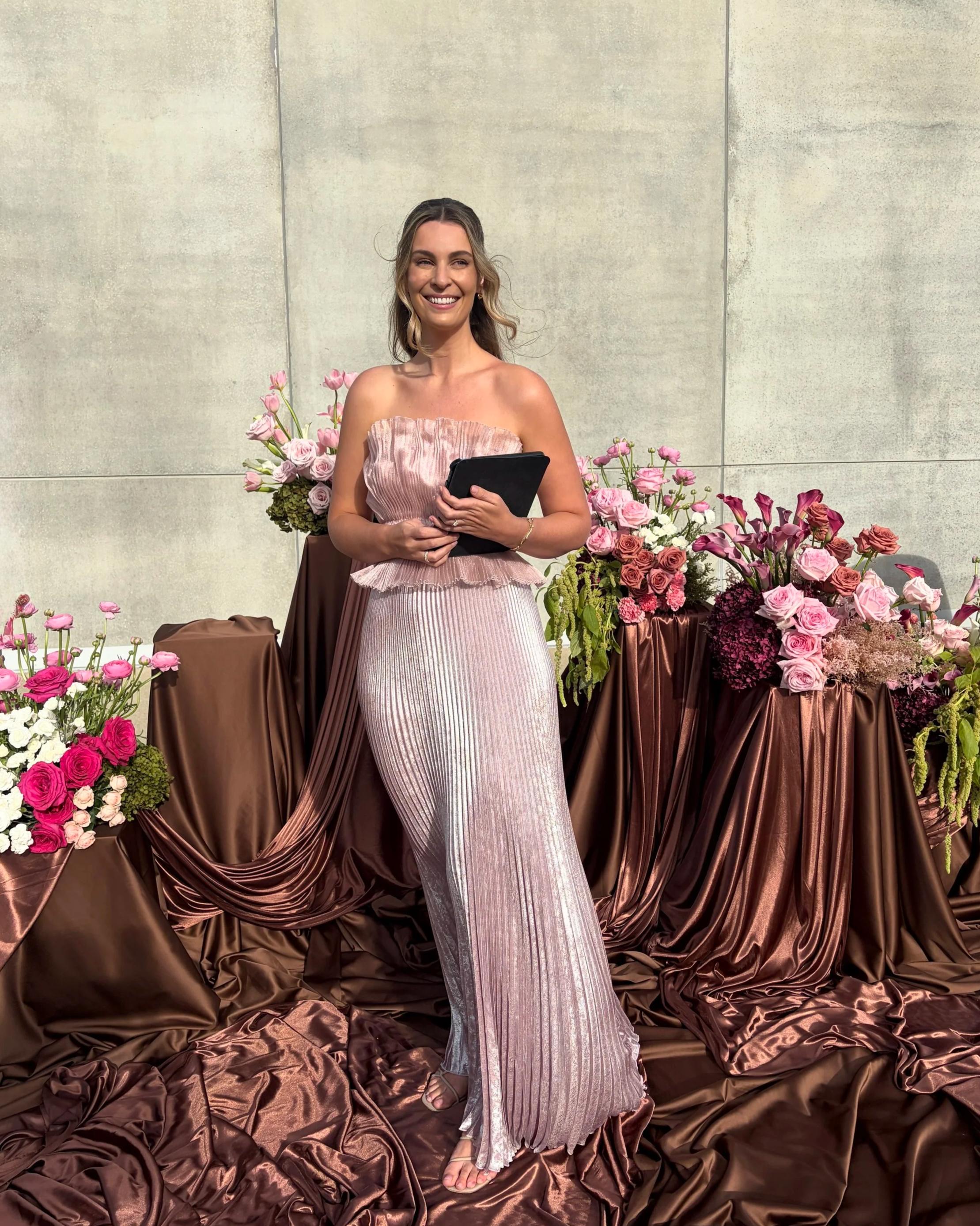 Love By Britt is standing in front of a wall with a flower display. She is smiling, wearing a strapless pink and silver pleated dress, holding a black card or tablet, and surrounded by pink and purple flowers on tables covered in brown satin fabric.