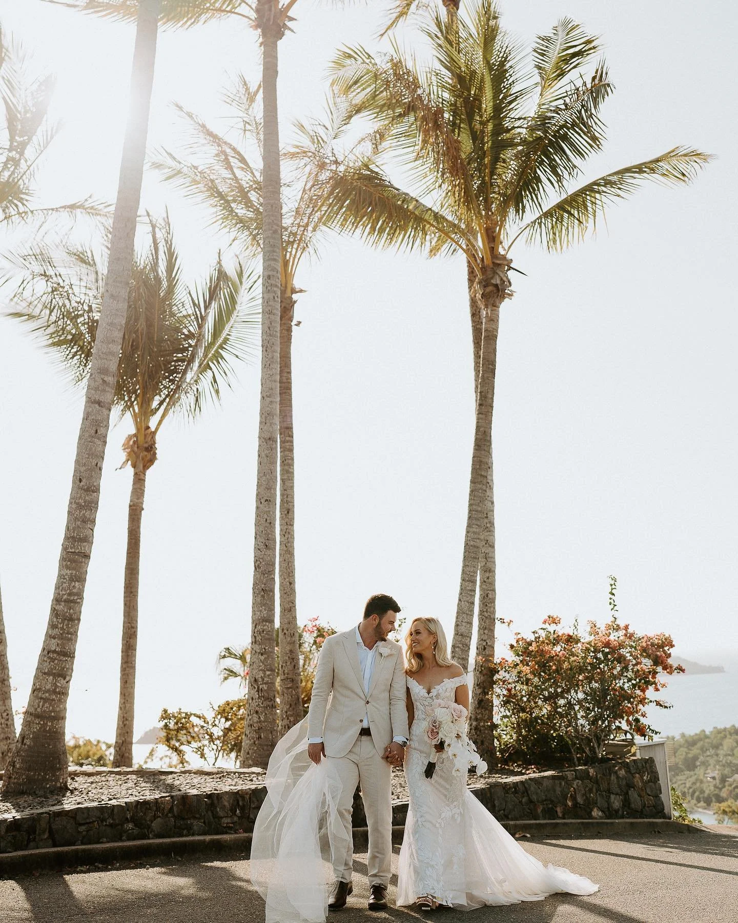 A bride and groom walking hand in hand outdoors near palm trees, with the bride holding a bouquet of flowers.