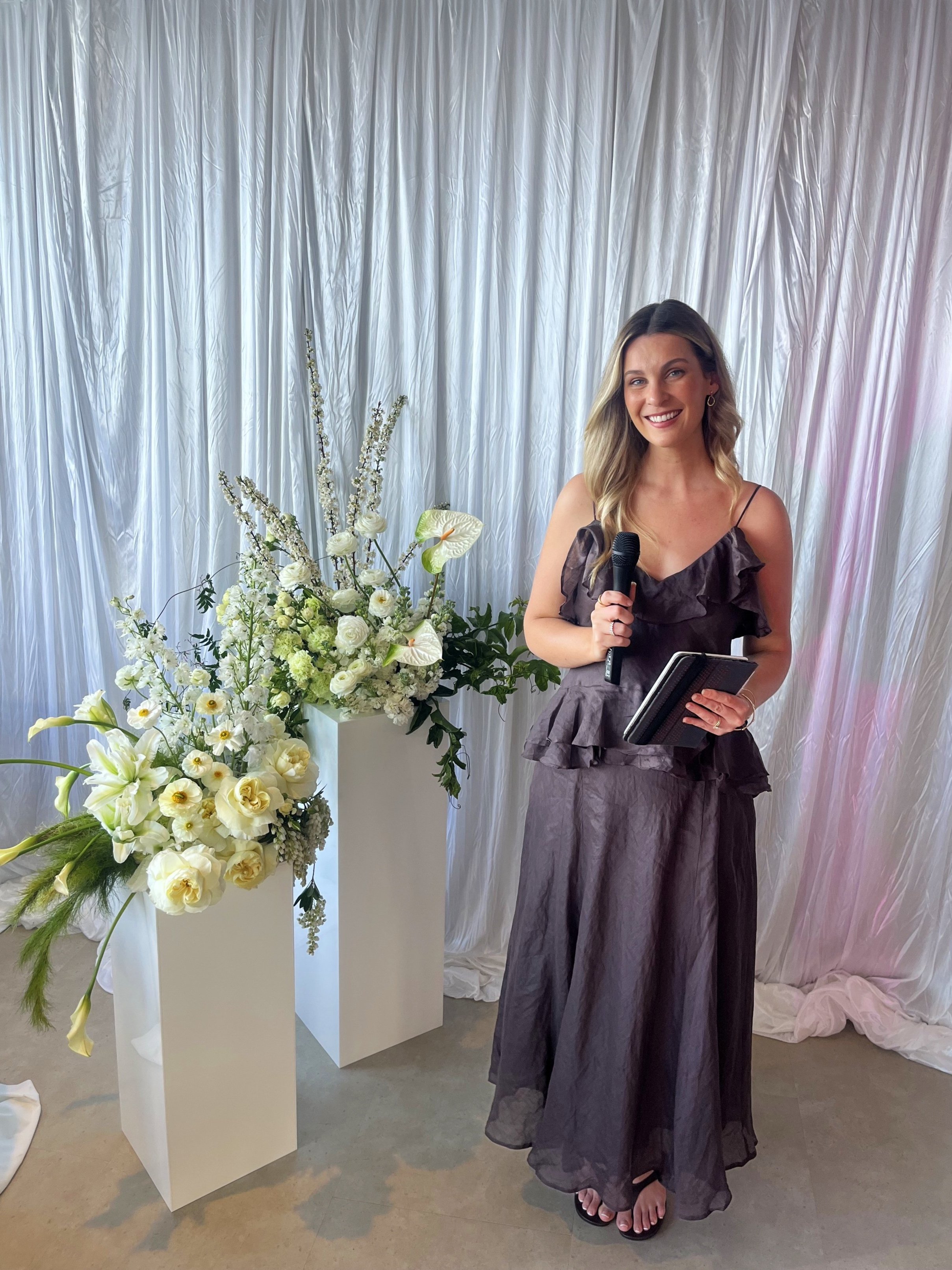 Brisbane Celebrant Love By Britt standing indoors, holding a microphone and a closed notebook, dressed in a dark ruffled dress, next to large white floral arrangements on white pedestals with a backdrop of white curtains.