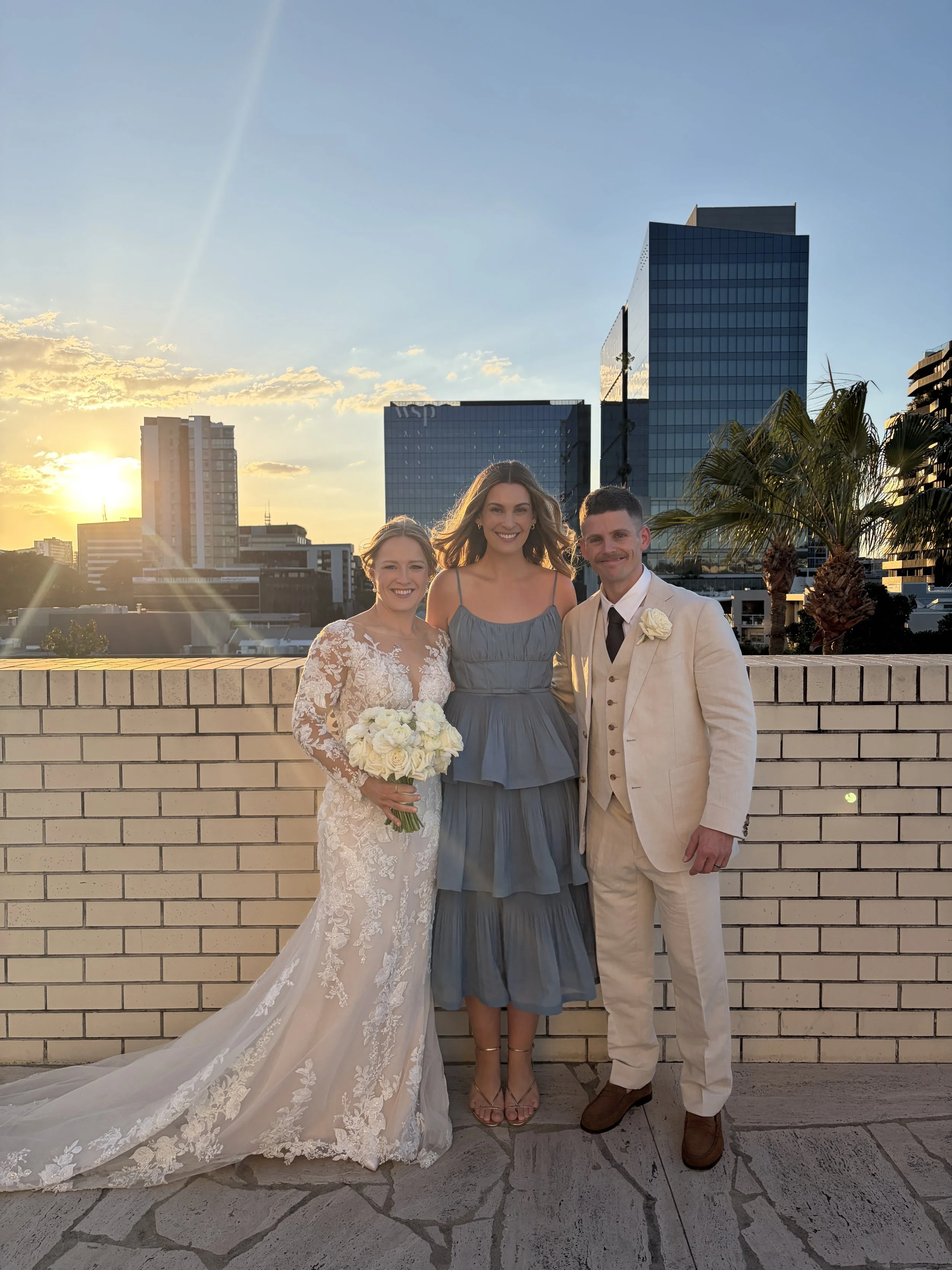 A bride in a white lace wedding dress holding a bouquet, a woman in a blue dress, and a man in a cream suit with a white shirt and tie, standing outside during sunset with a cityscape background of tall modern buildings and palm trees.