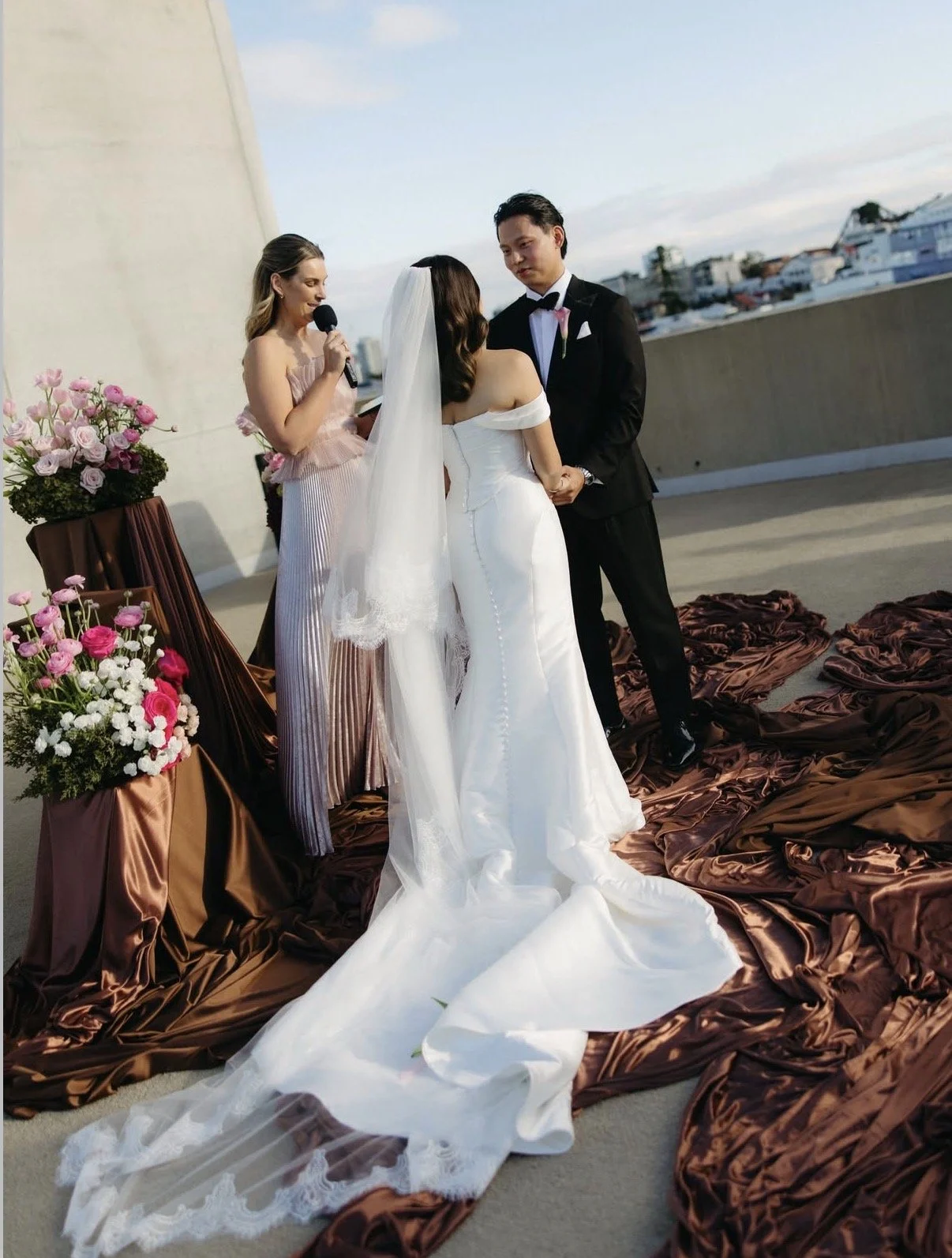 A wedding ceremony on a rooftop with a bride and groom holding hands, Britt is conducting their ceremony, pink roses, and brown velvet drapes on the floor.