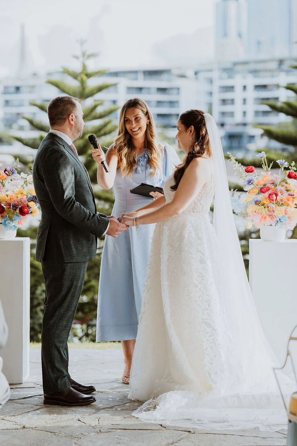 A wedding ceremony with a bride and groom holding hands and facing each other, while Love By Britt Celebrant stands between them smiling and holding a microphone. and trees.
