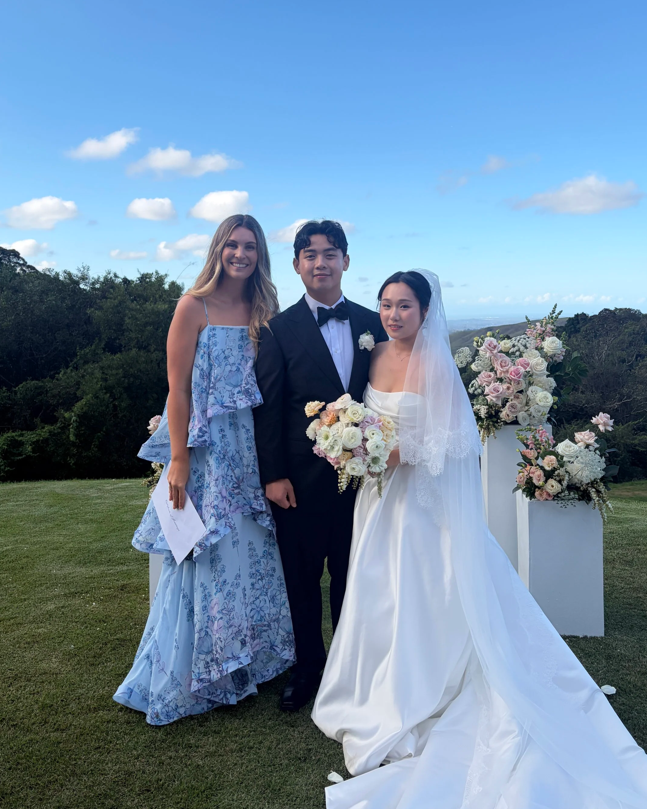 A group of three people at a wedding outdoors, with two women and one man. One woman is wearing a light blue dress, the man in a black tuxedo, and the other woman in a white wedding gown with a veil. They are standing on a grassy area with trees and flowers around them, under a blue sky with scattered clouds.