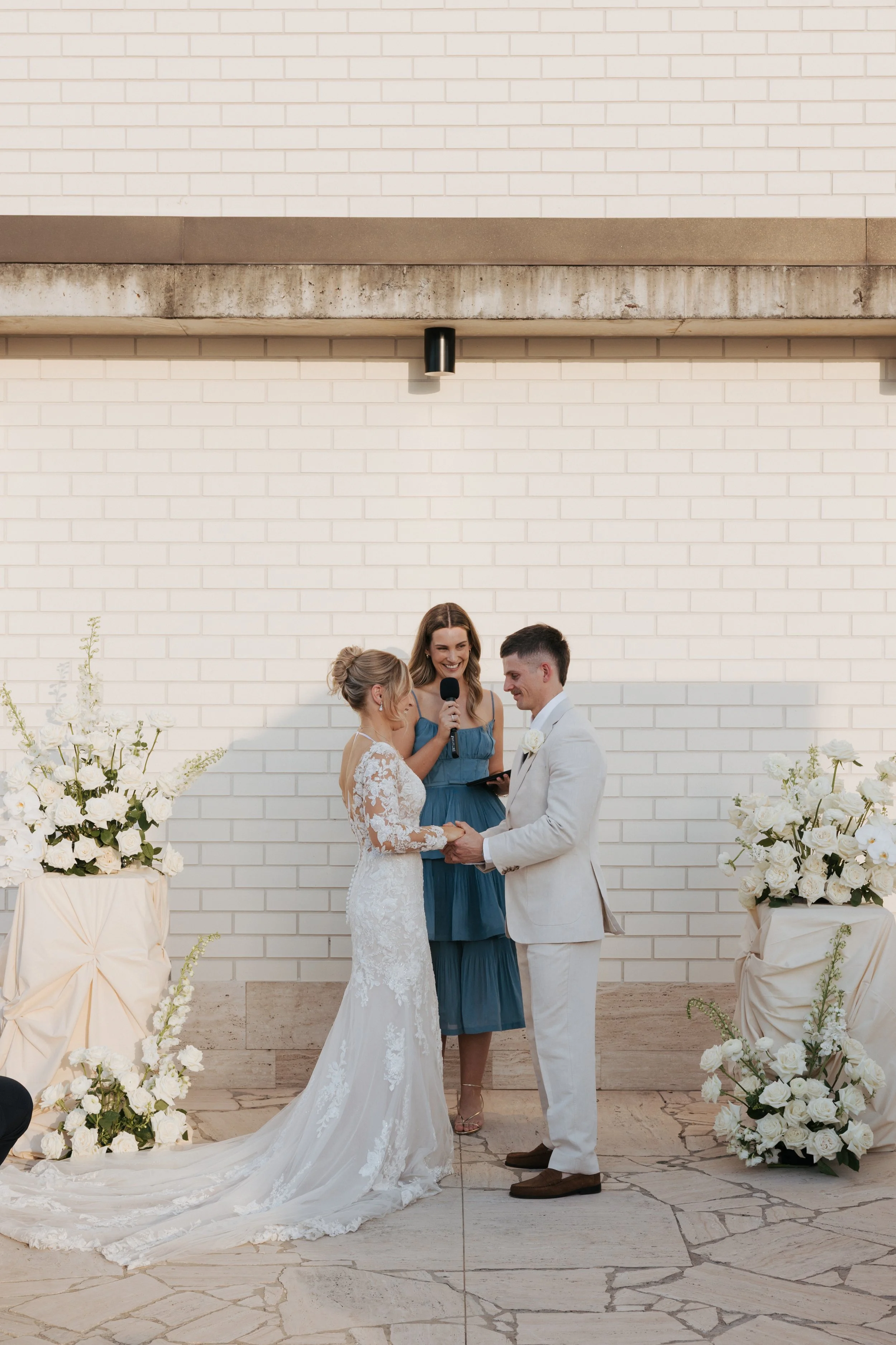 A couple getting married, holding hands and exchanging vows, with Love By Britt Celebrant standing between them holding a microphone in front of a white brick wall with floral arrangements on either side.