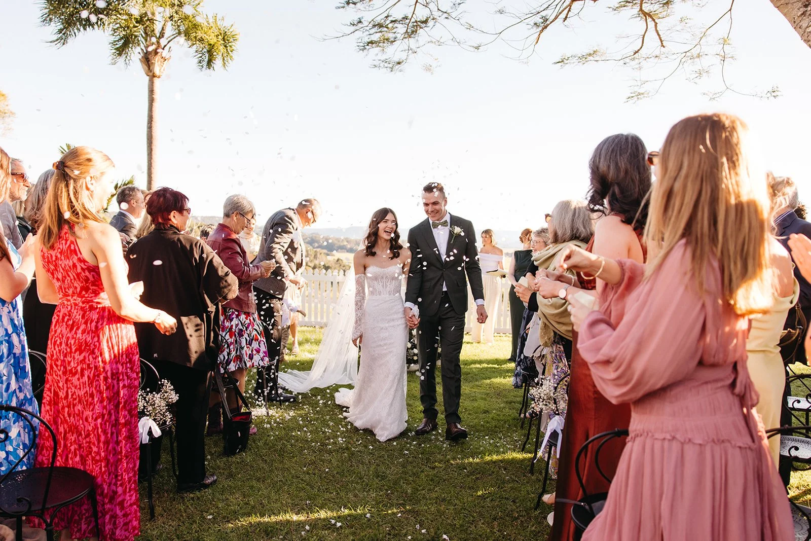 A newlywed couple walking hand in hand down an outdoor aisle, surrounded by celebrating guests on a sunny day with trees and a white picket fence in the background.