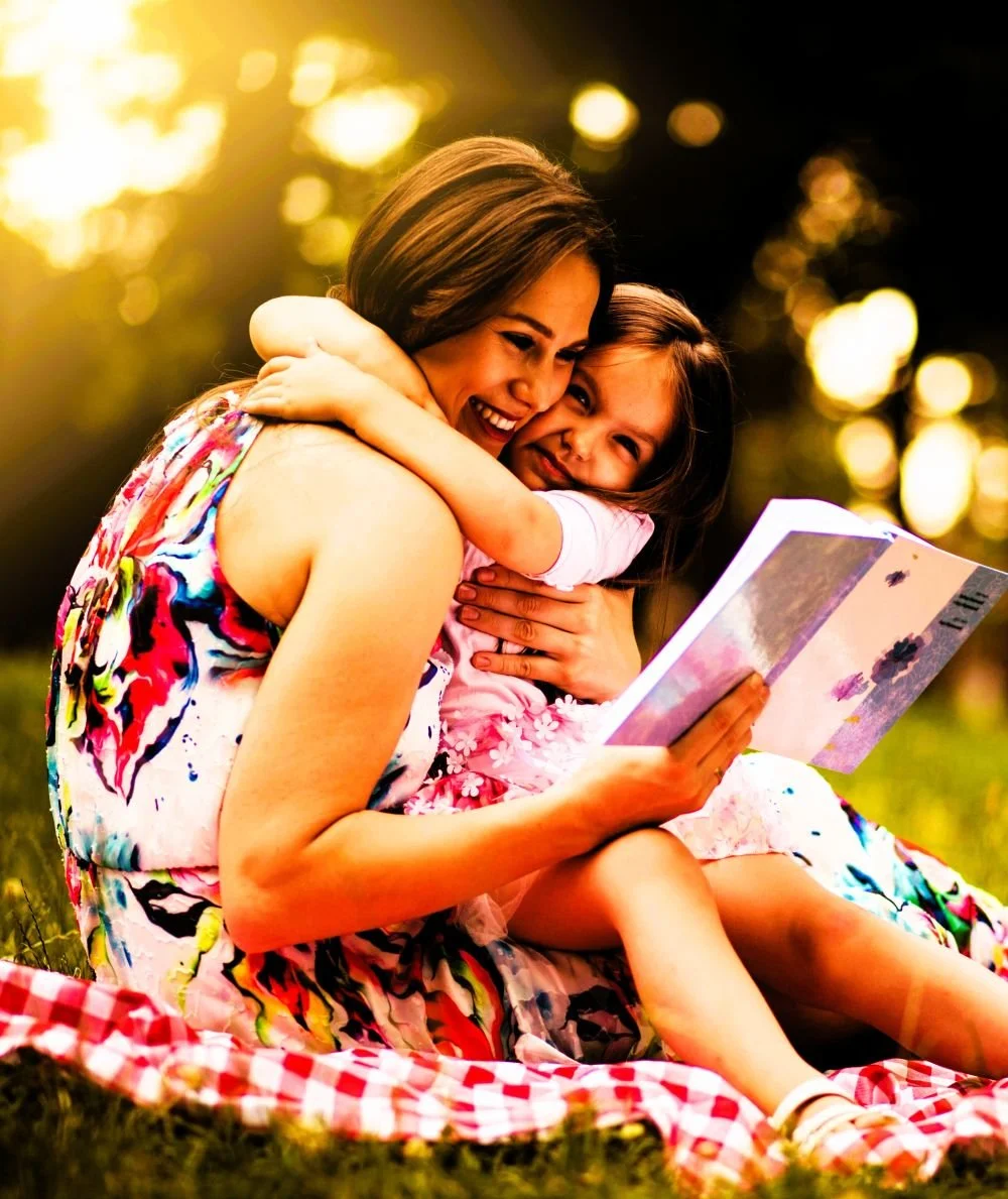 mother hugging daughter while reading outside on a picnic blanket