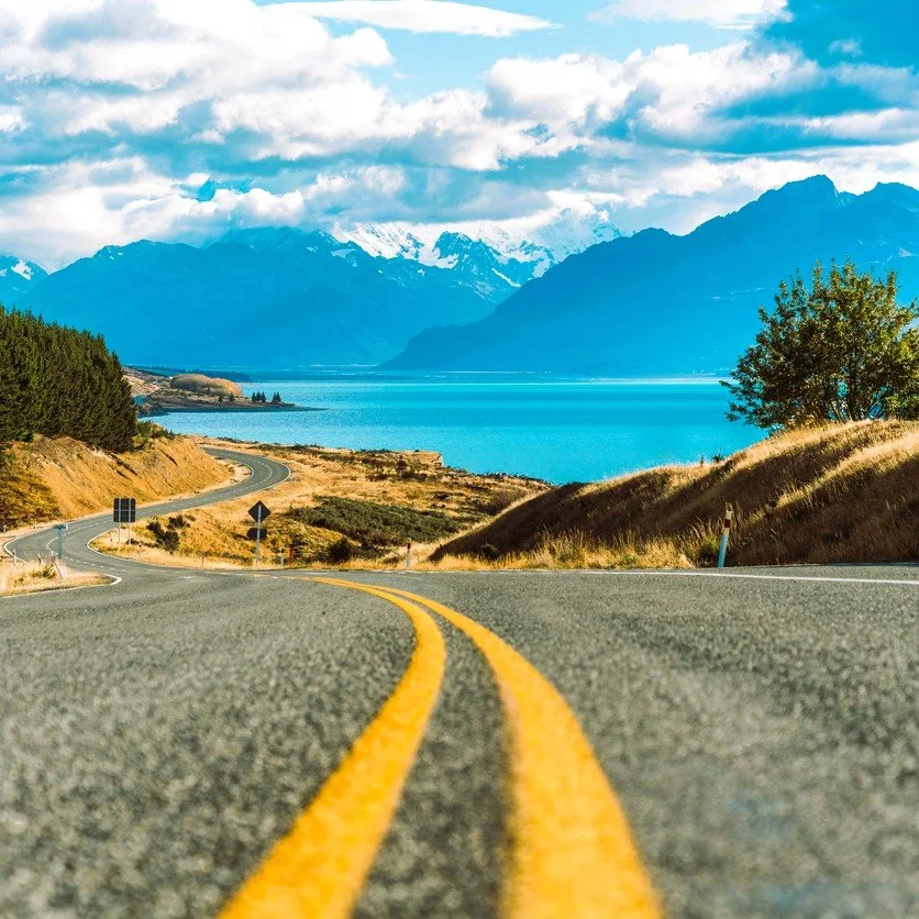 image of road curving into the distance with a lake and mountains in the background