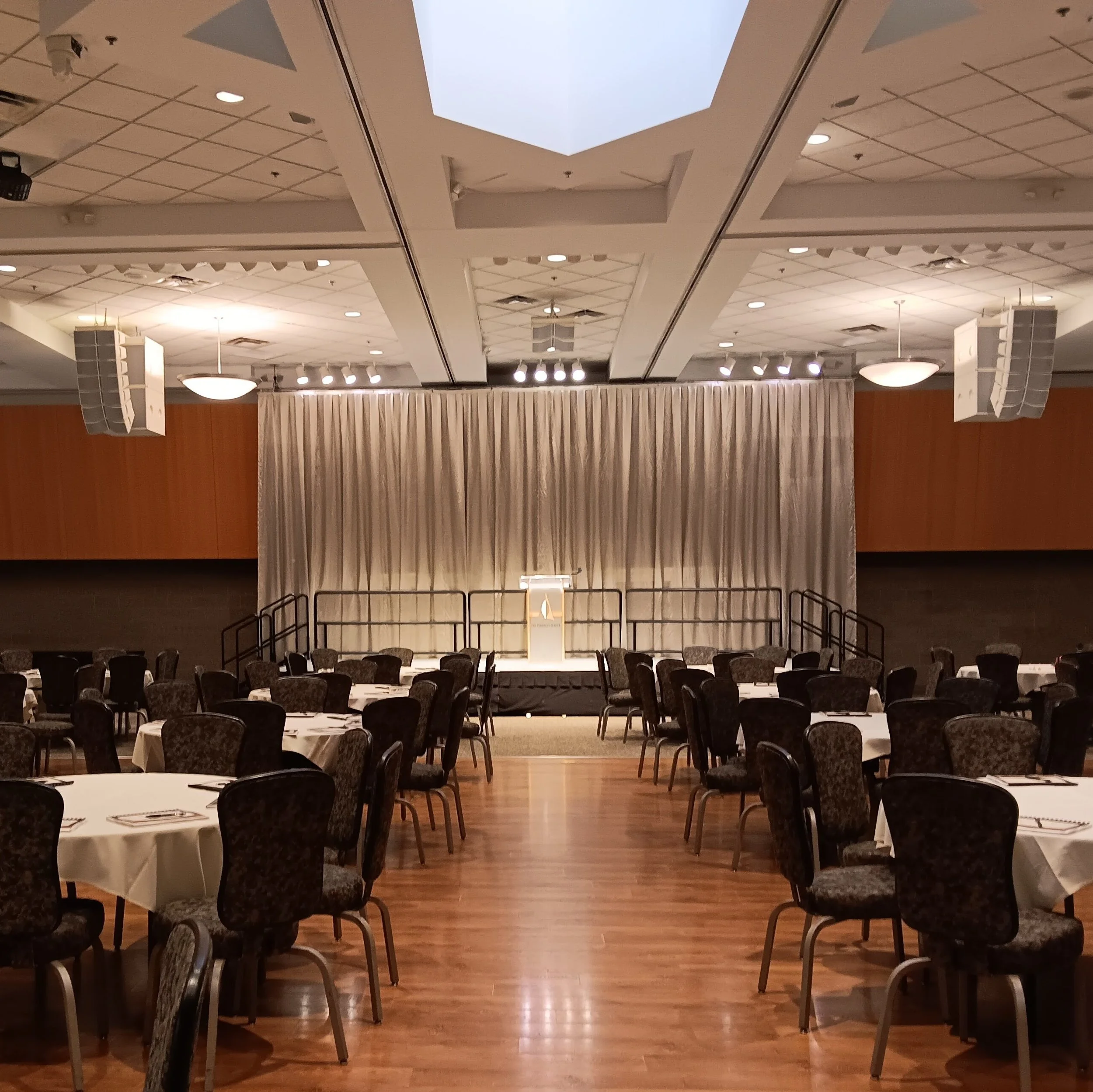 Empty banquet hall with round tables covered in white tablecloths, set with utensils, and chairs arranged around each table. In the background, a stage with a podium and curtained backdrop. Overhead lighting and speakers visible in the ceiling.