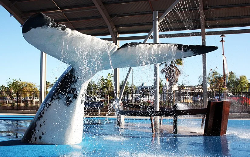 Whale-shaped water fountain in a pool with water spraying from its blowhole, surrounded by a shaded area and outdoor scenery.