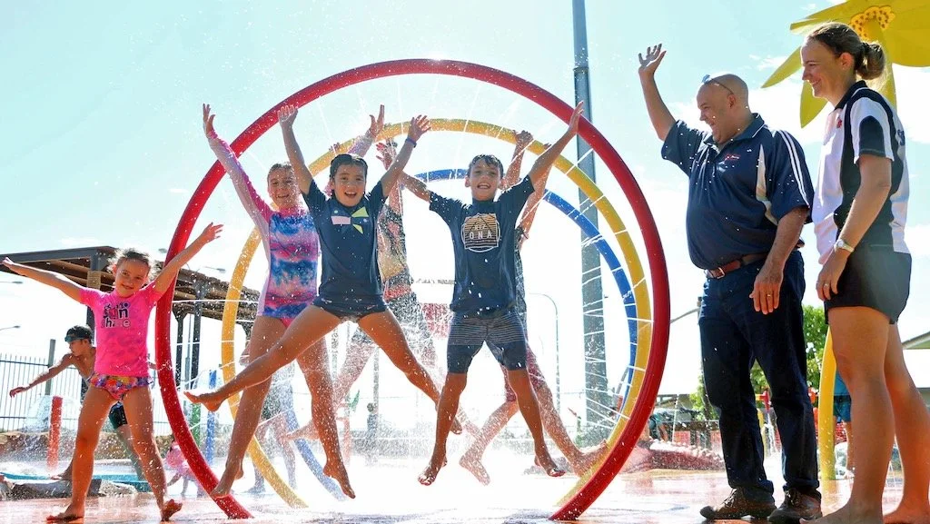 Children playing and splashing water in a colorful splash pad, with two adults watching and smiling nearby.