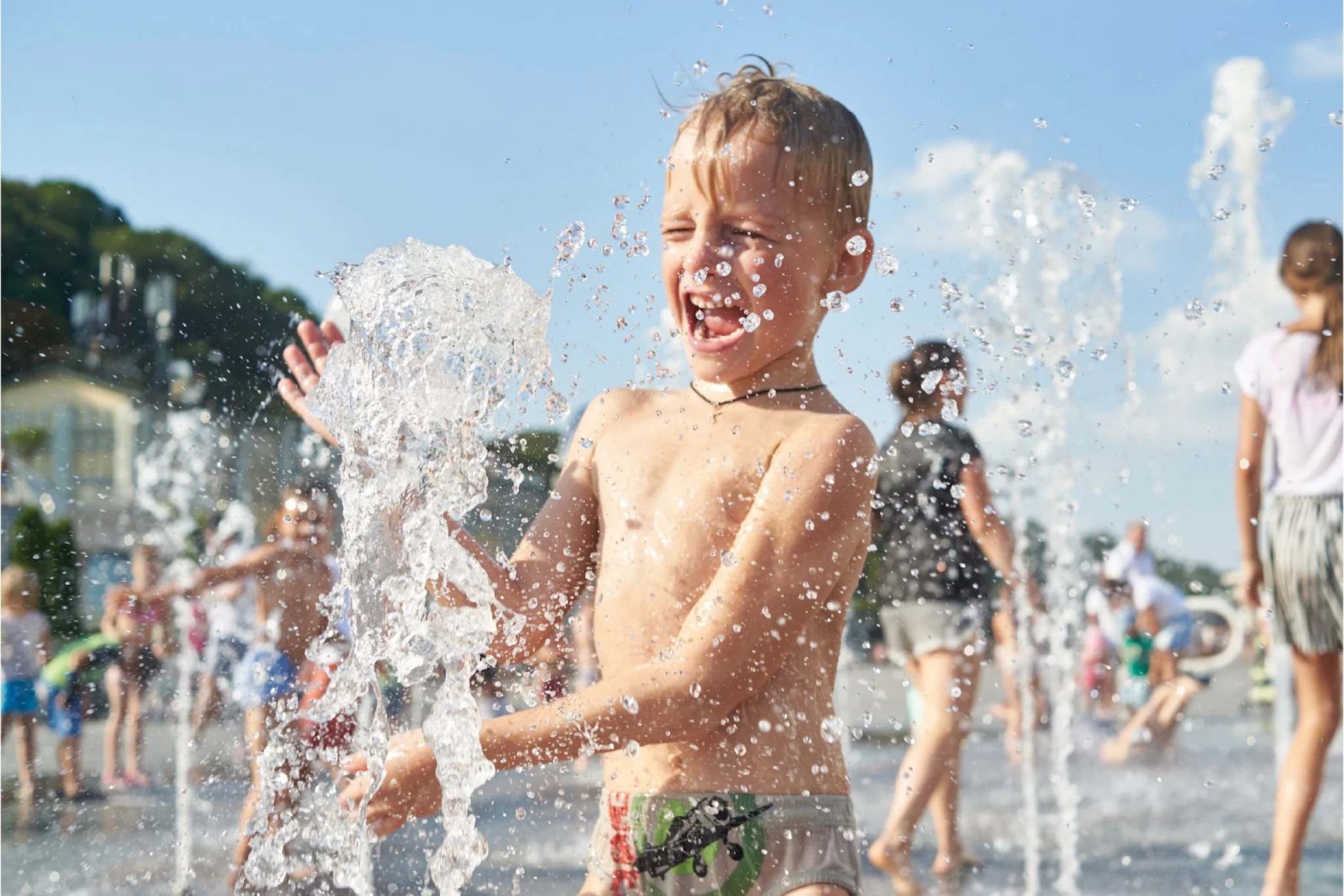 A young boy playing in water at a splash pad or fountain, smiling and splashing water on a sunny day, with other kids playing in the background.