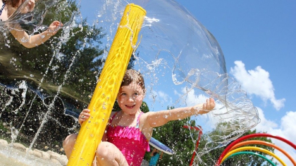 A young girl in a pink swimsuit playing with a water toy outdoors on a sunny day, surrounded by spray and colorful hoops.