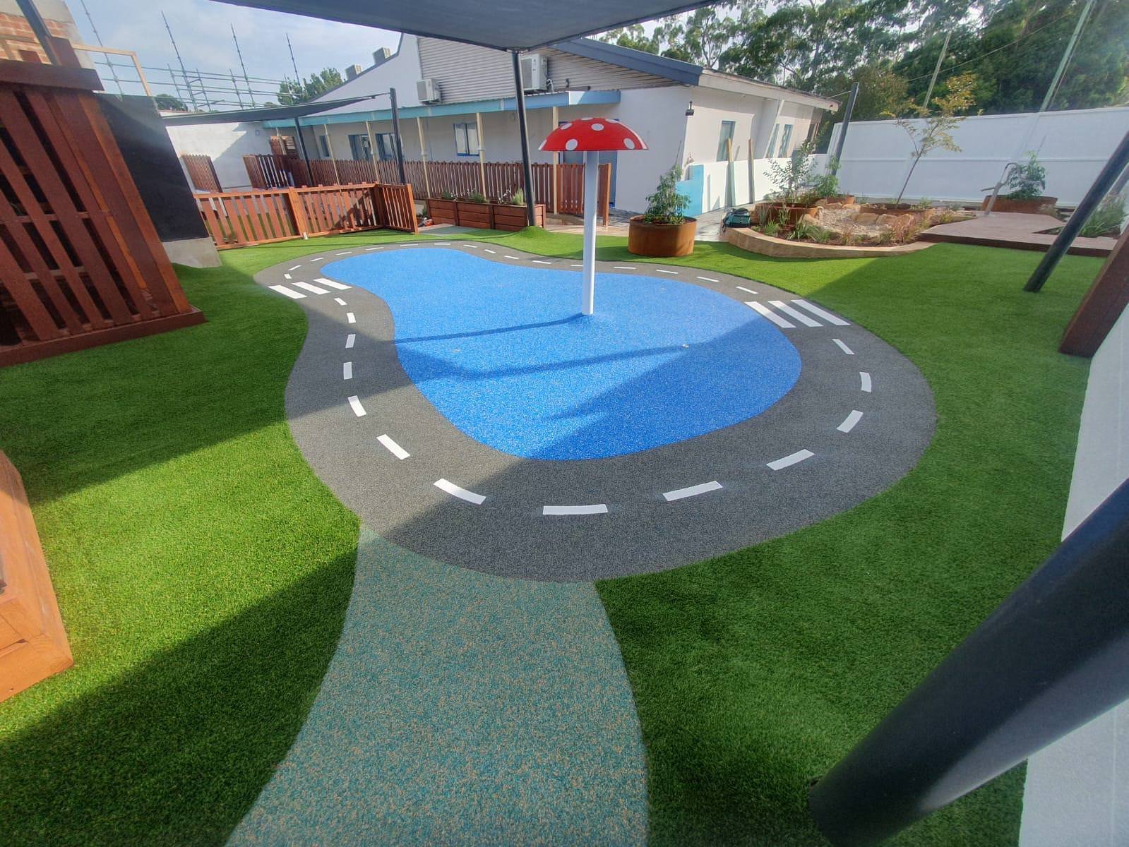 An outdoor play area for children featuring a small blue splash pad with a red and white mushroom-shaped umbrella, surrounded by artificial grass and a curved pathway.