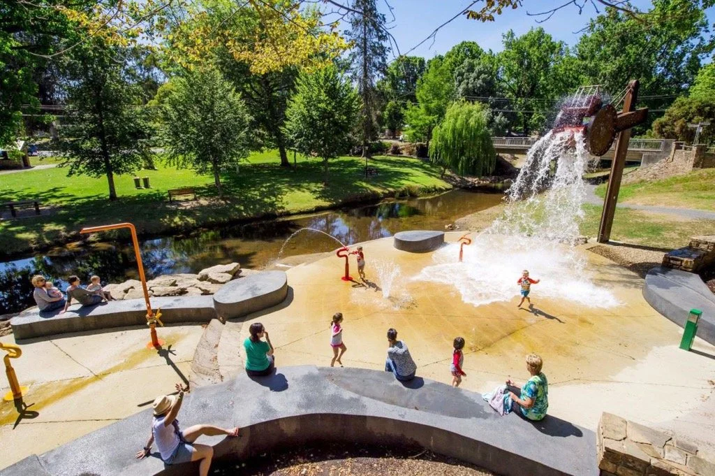 Children playing in a splash pad with a water wheel and water spraying from a pipe, surrounded by trees and park visitors.