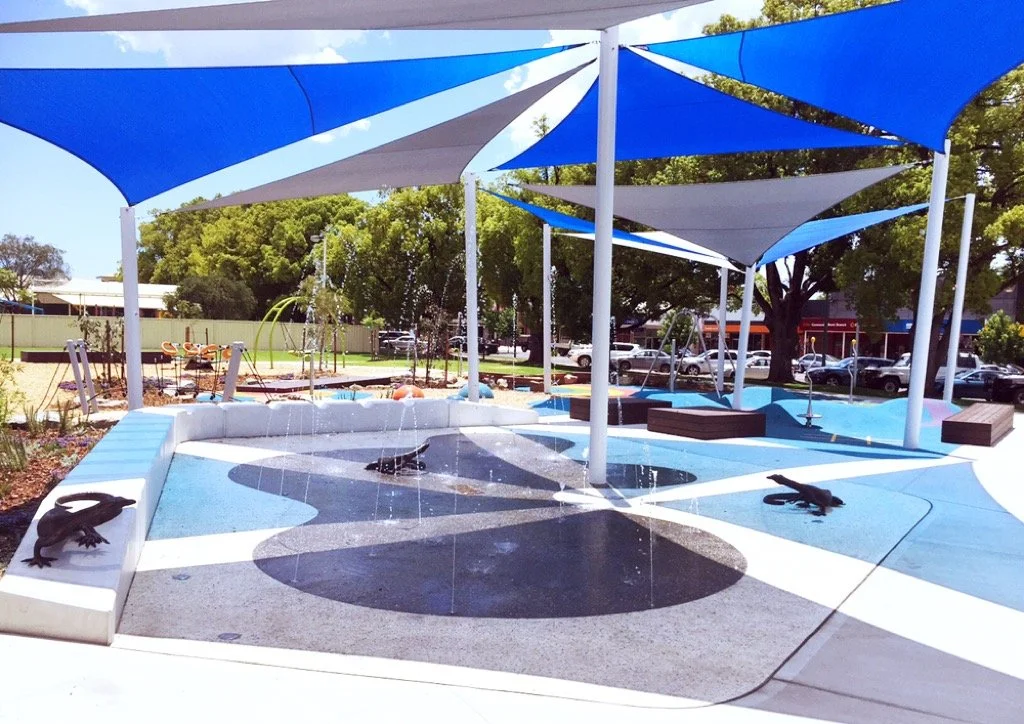 Outdoor splash pad with water features, shaded by blue and white fabric canopies, surrounded by trees and parked cars.