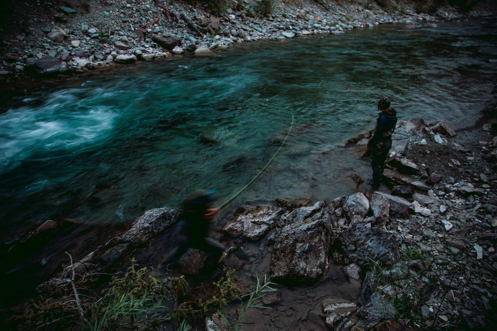 A person standing on rocky riverbank holding a fishing rod in a flowing river