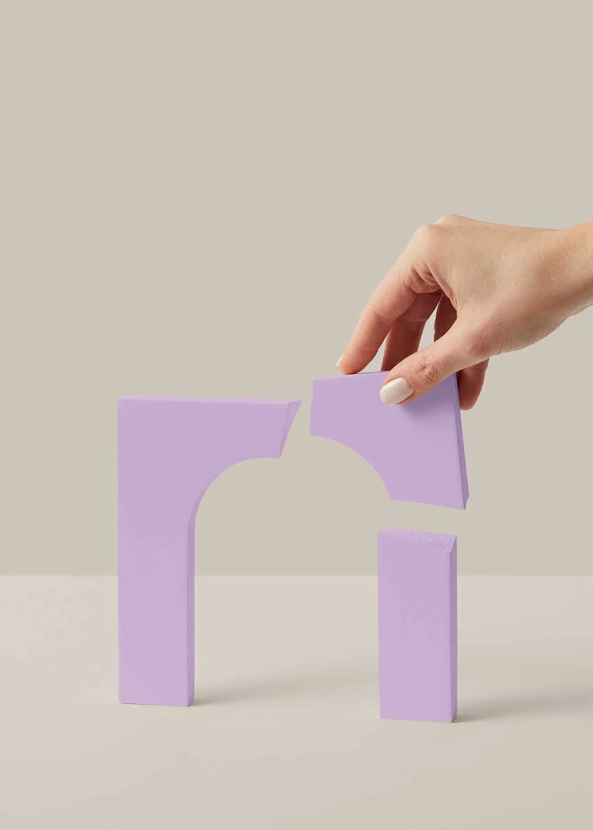 A hand stacking two lilac-colored foam blocks to create an arch shape. The blocks have smooth surfaces and are arranged on a beige surface with a plain light background.