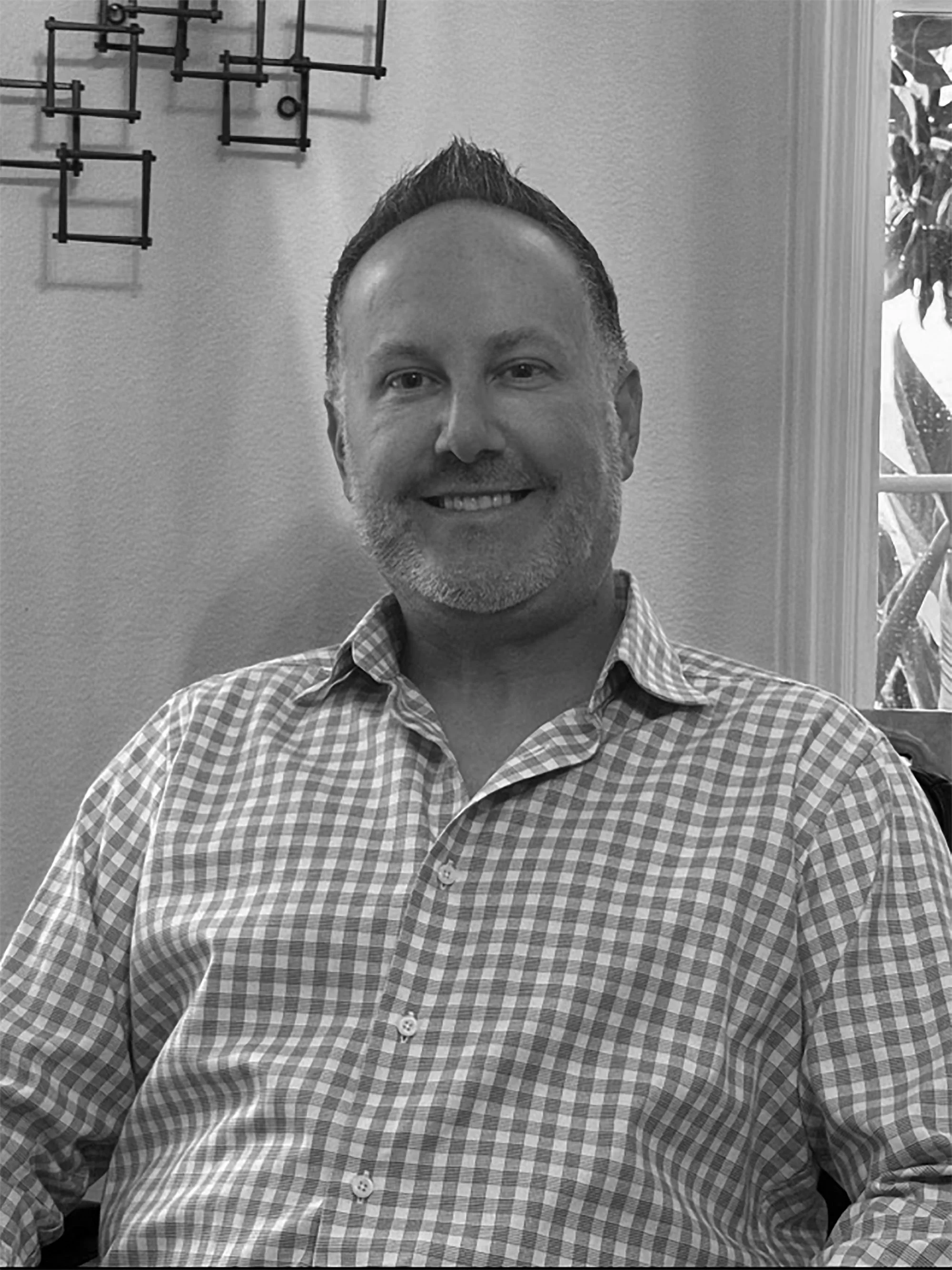 A man with short hair and a beard smiling, wearing a checkered shirt, sitting indoors, with decorative wall art and a window with a view of trees