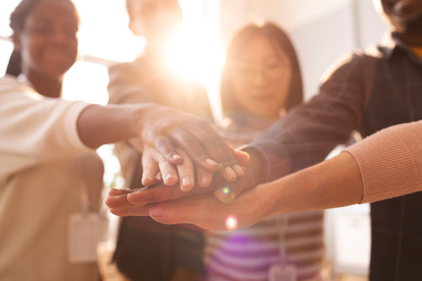 Diverse hands stacked together with bright sunlight in the background, symbolizing teamwork.