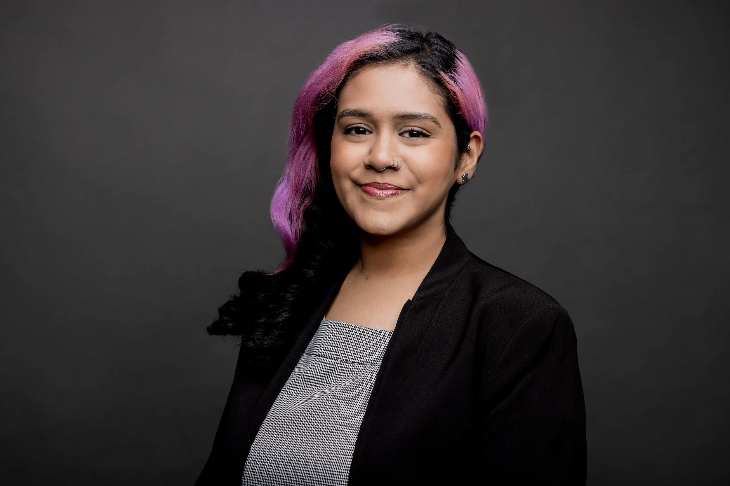Young woman with pink and black hair, wearing a blazer, smiling against a dark background.