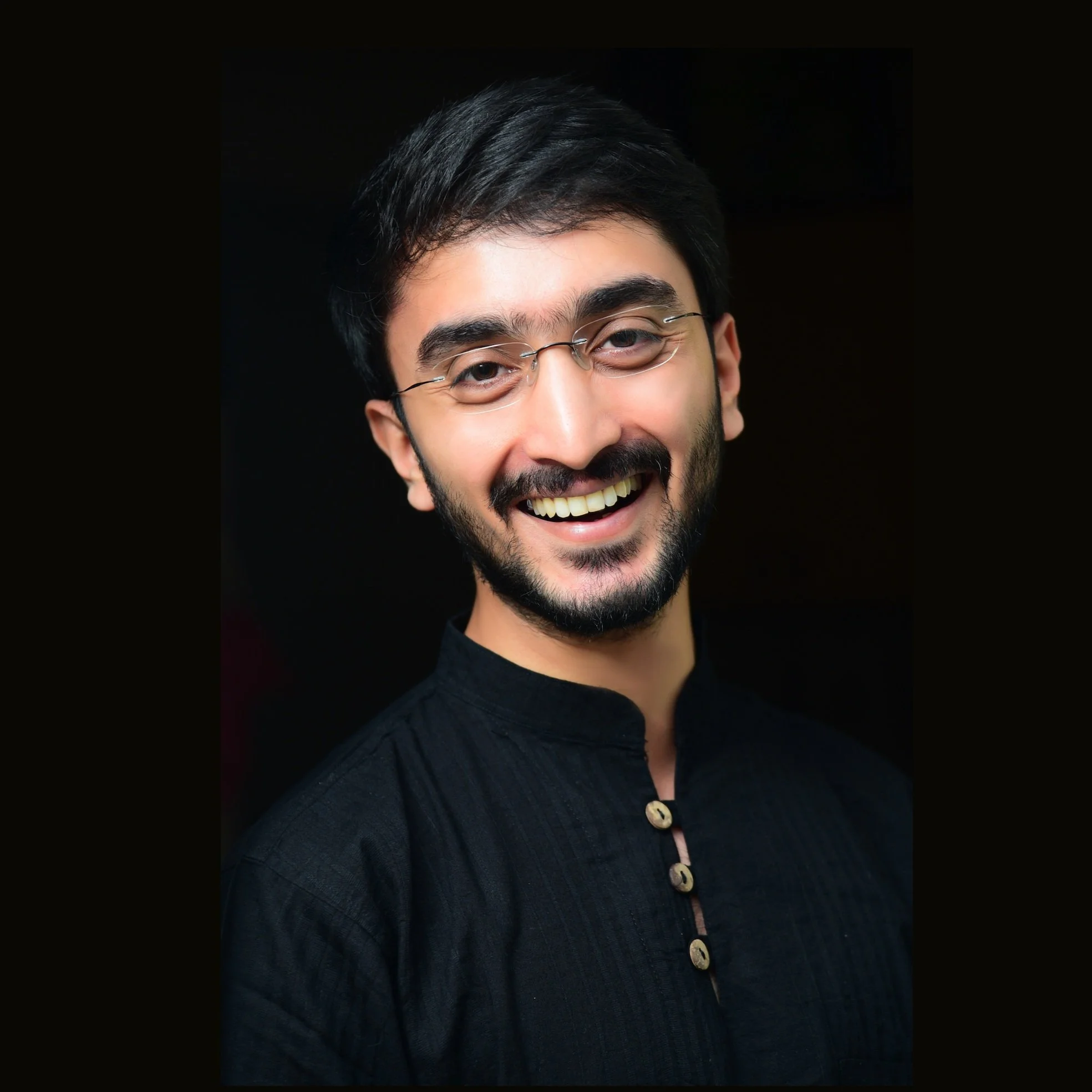 Man with glasses and a beard smiles warmly, wearing a black kurta against a dark background.