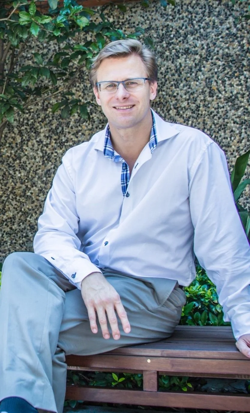Man sitting on a wooden bench, wearing a light shirt and smiling against a textured backdrop.