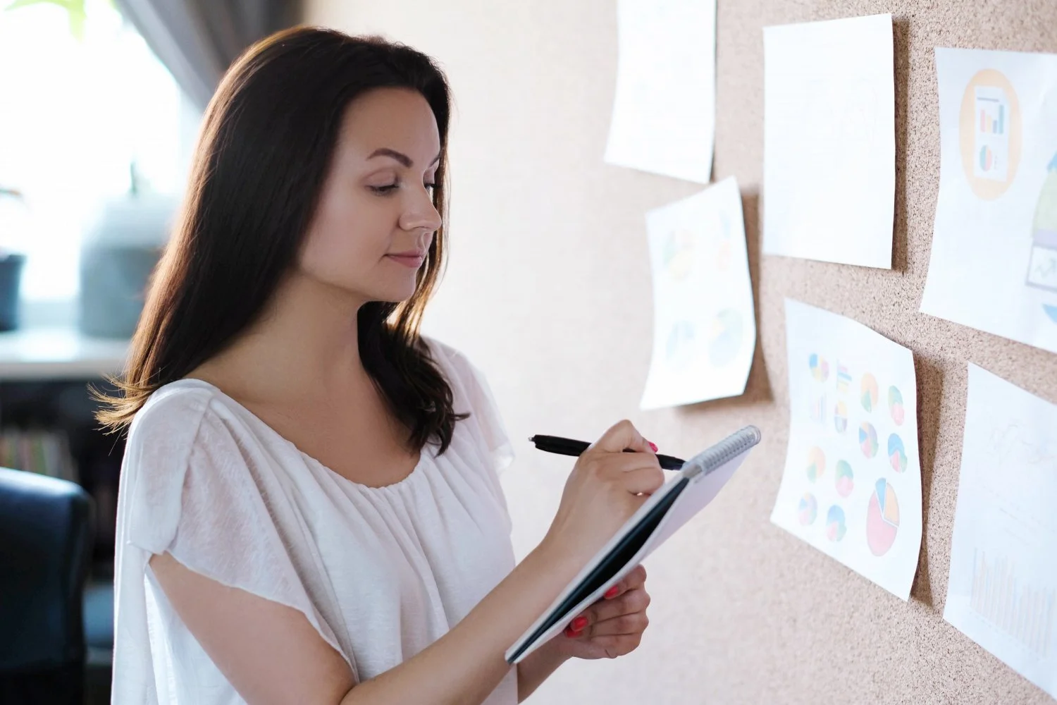 Woman with long dark hair writes in a notebook while examining charts on a corkboard.