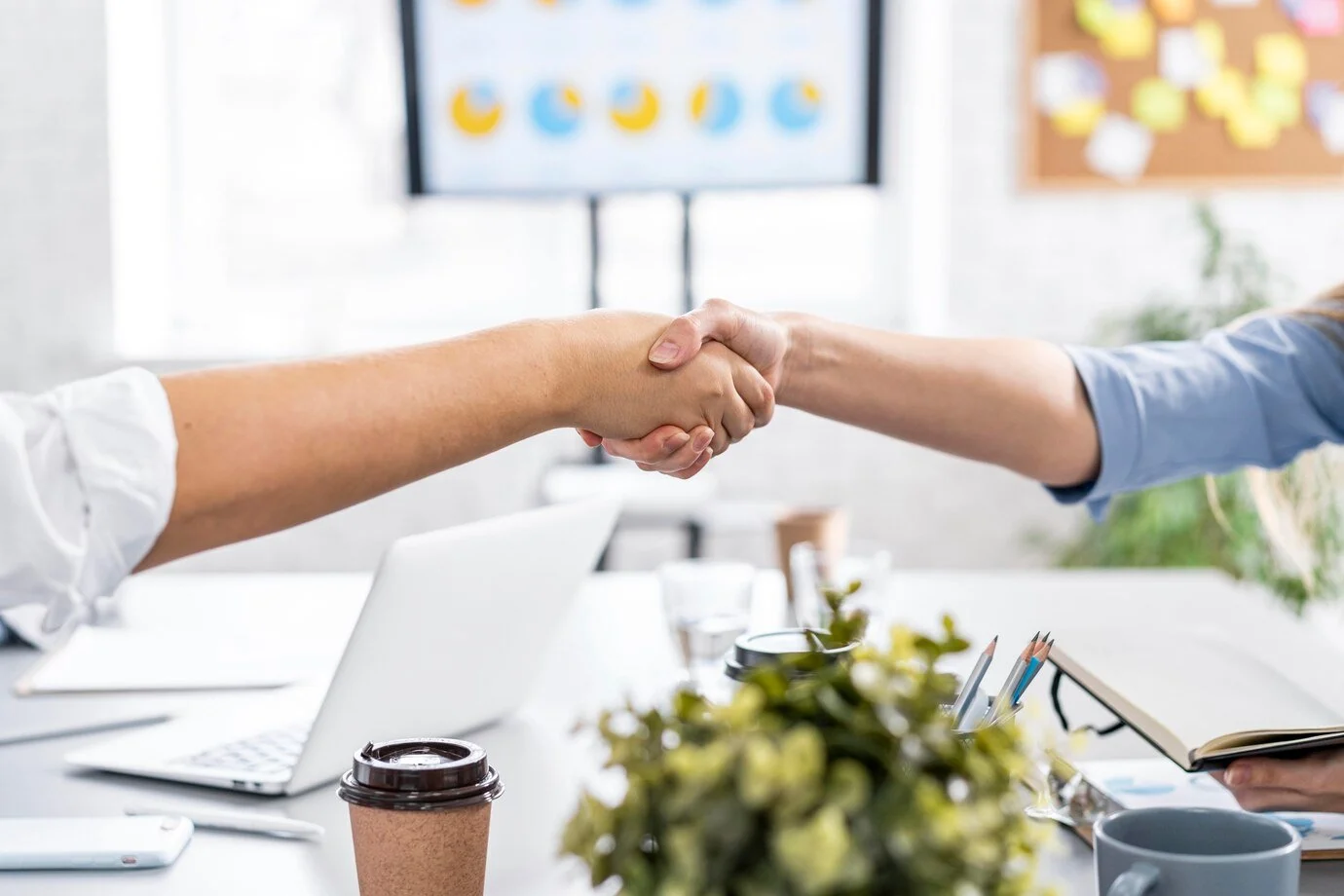 Two individuals shaking hands over a desk with a laptop and coffee cup in the foreground.