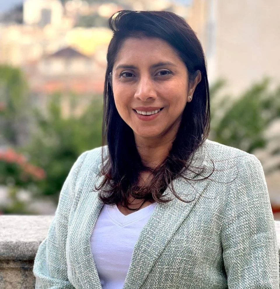 Smiling woman in a light green blazer stands outdoors with blurred background.