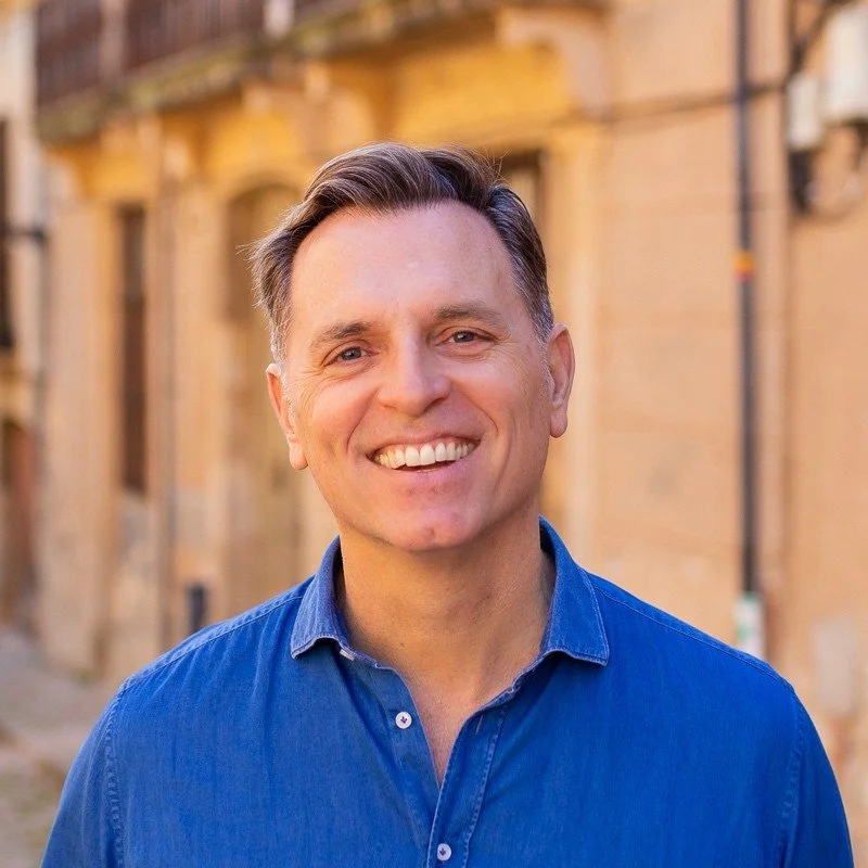 Smiling man in a blue shirt stands outdoors with a historic building in the background.