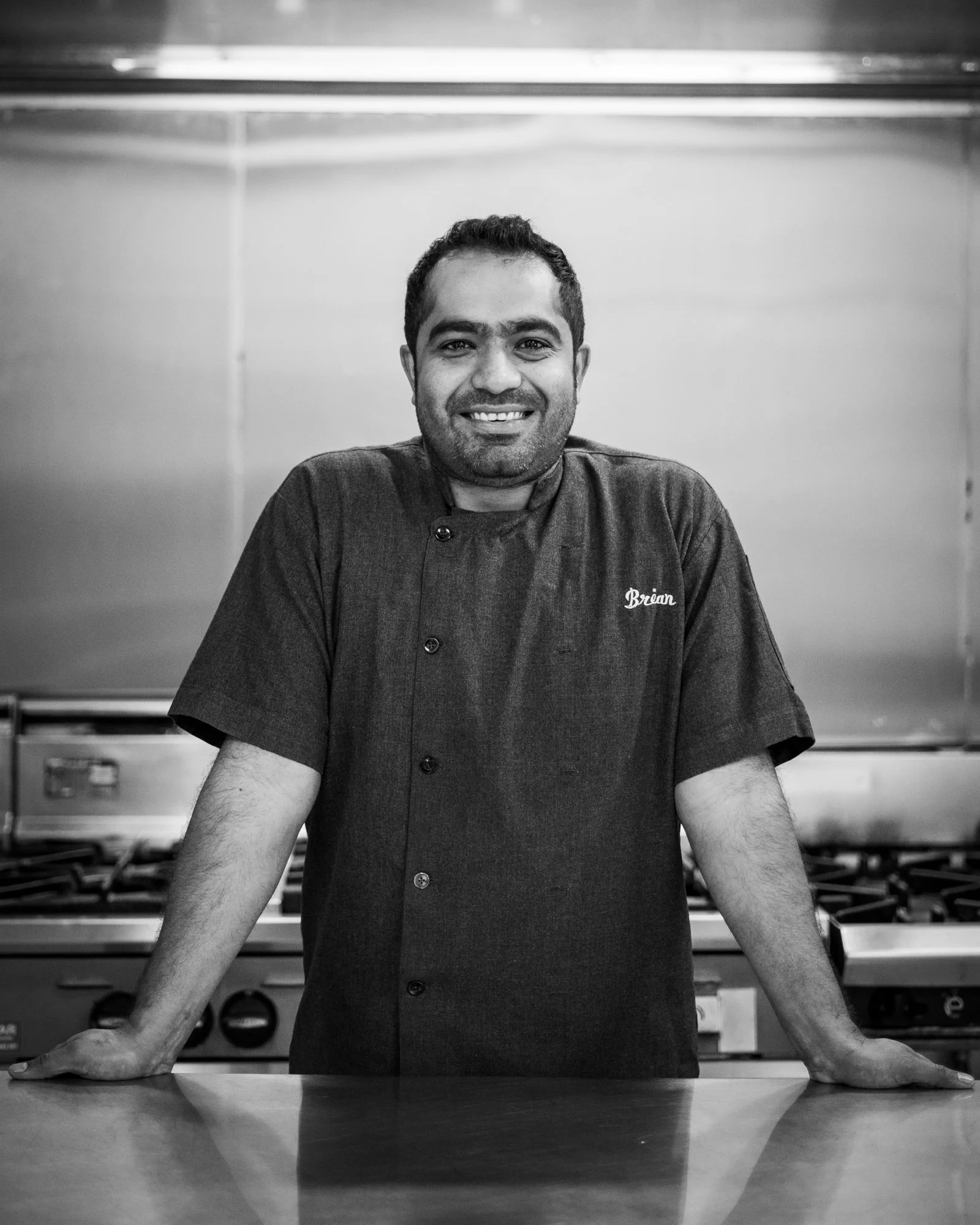 Man in a chef's coat standing in a professional kitchen with a stove in the background.