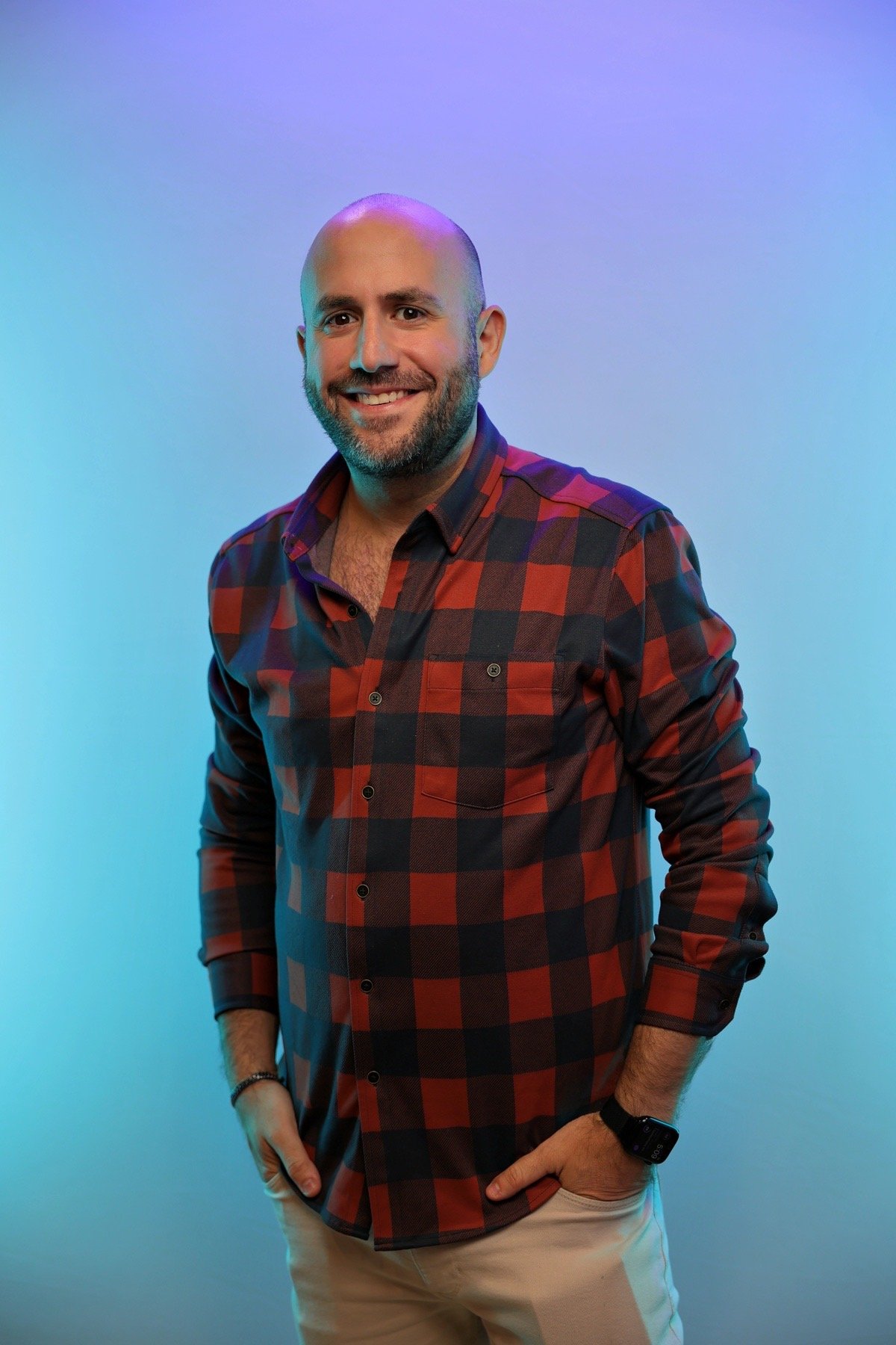 A smiling man with a beard and a shaved head, wearing a red and black plaid shirt, beige pants, and a smartwatch, standing against a blue and purple gradient background.
