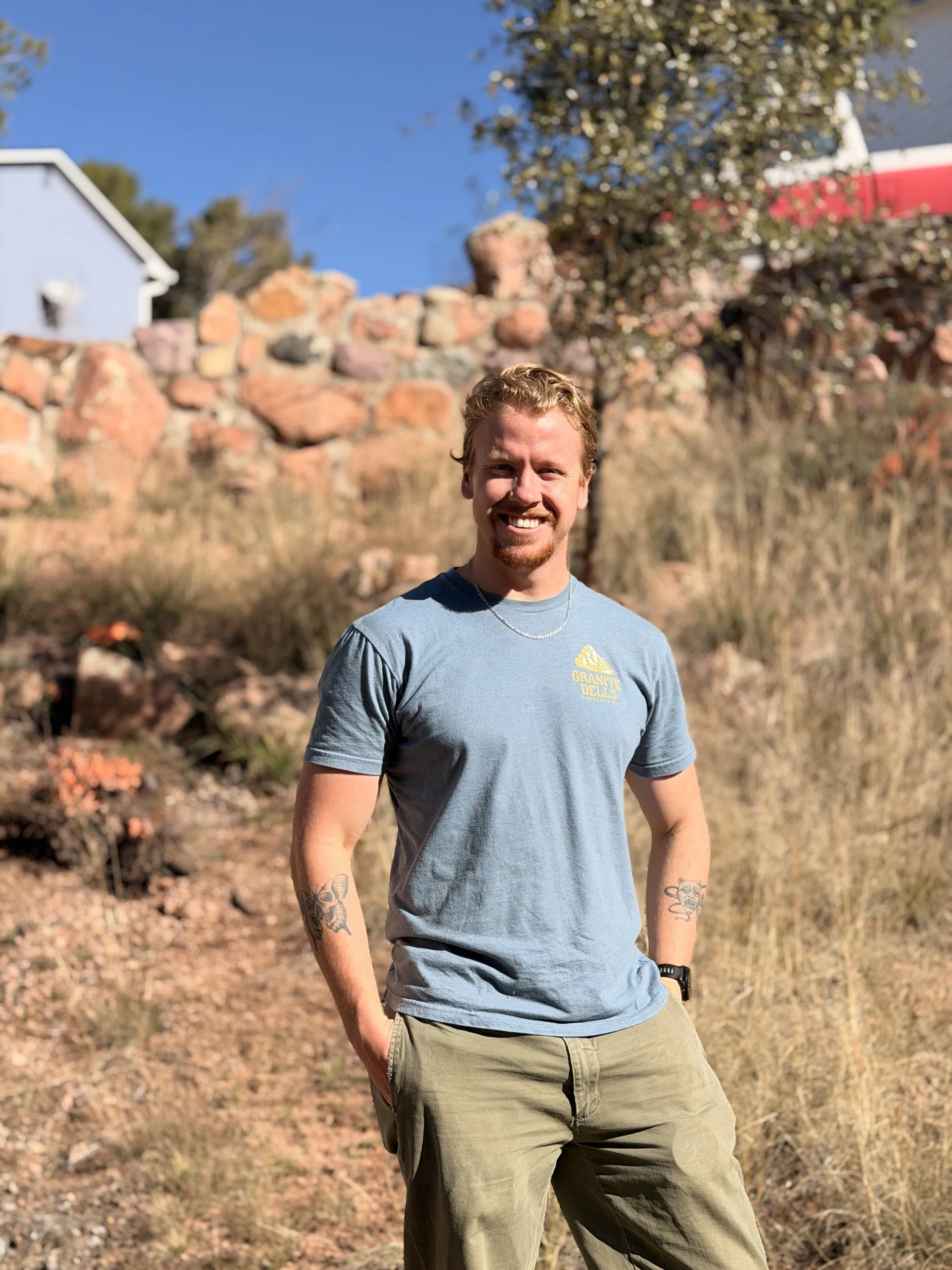 A young man with curly blonde hair and a beard, smiling, wearing a light blue T-shirt and khaki pants, standing outdoors with a rocky background, dry grass, and a house with a red roof in the distance on a sunny day.