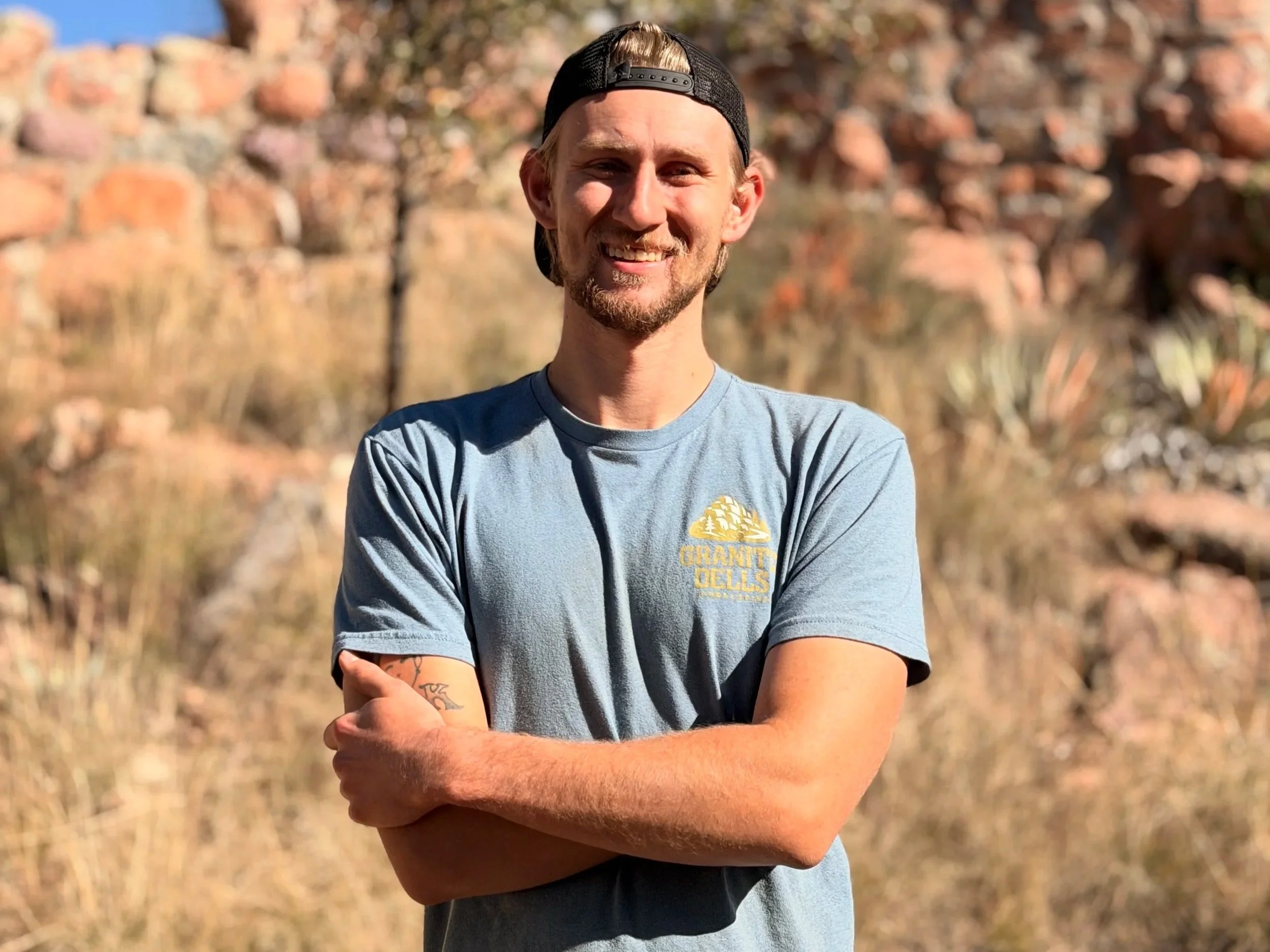 A young man with a beard and blonde hair, wearing a backwards black baseball cap and a light baseball-style T-shirt with a graphic and text, smiling with arms crossed, outdoors with desert and rocks background.