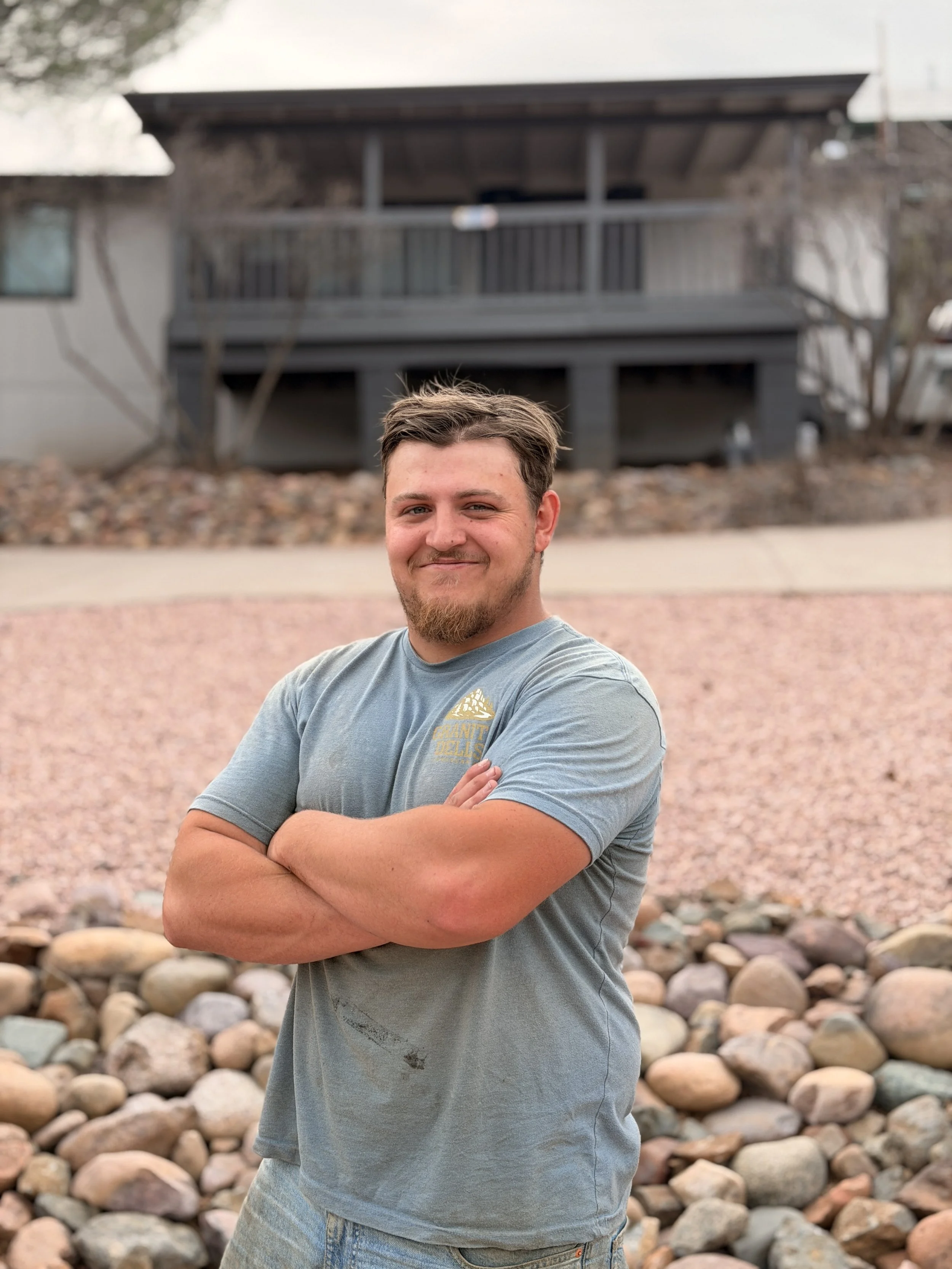 A young man with a beard and short brown hair standing outdoors with arms crossed, smiling at the camera, in front of a rocky landscape and a dark gray house with a porch.