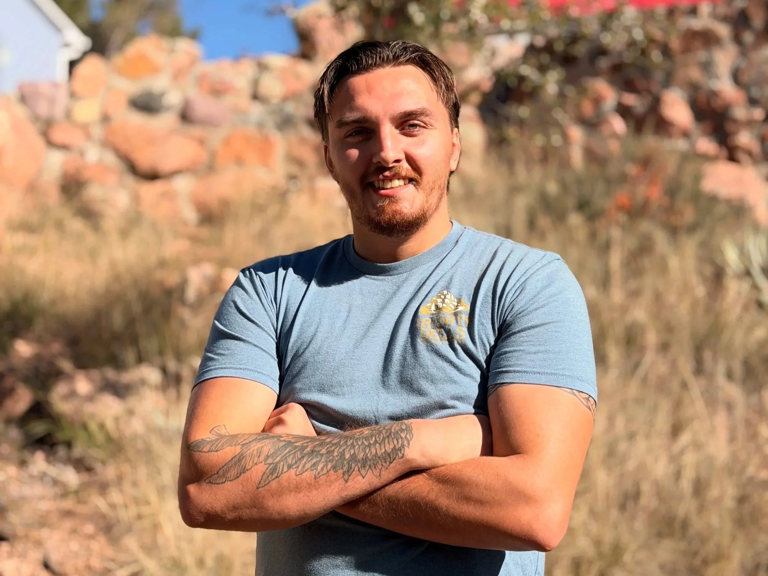 A young man with brown hair and a beard smiling, standing outdoors with arms crossed. He is wearing a light blue T-shirt with a logo on the chest, in front of a background of rocks and desert vegetation under clear sky.