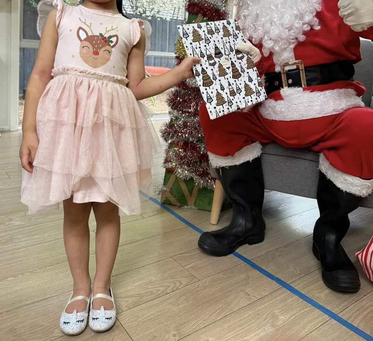 A young girl in a pink dress with a reindeer design, standing next to Santa Claus, who is sitting on a gray chair. Santa is dressed in his red suit with white fur trim, black boots, and a belt. The girl is holding a Christmas gift bag decorated with Christmas trees and reindeer patterns. Behind them is a small decorated Christmas tree with red tinsel and lights, and a glass door leading outside.