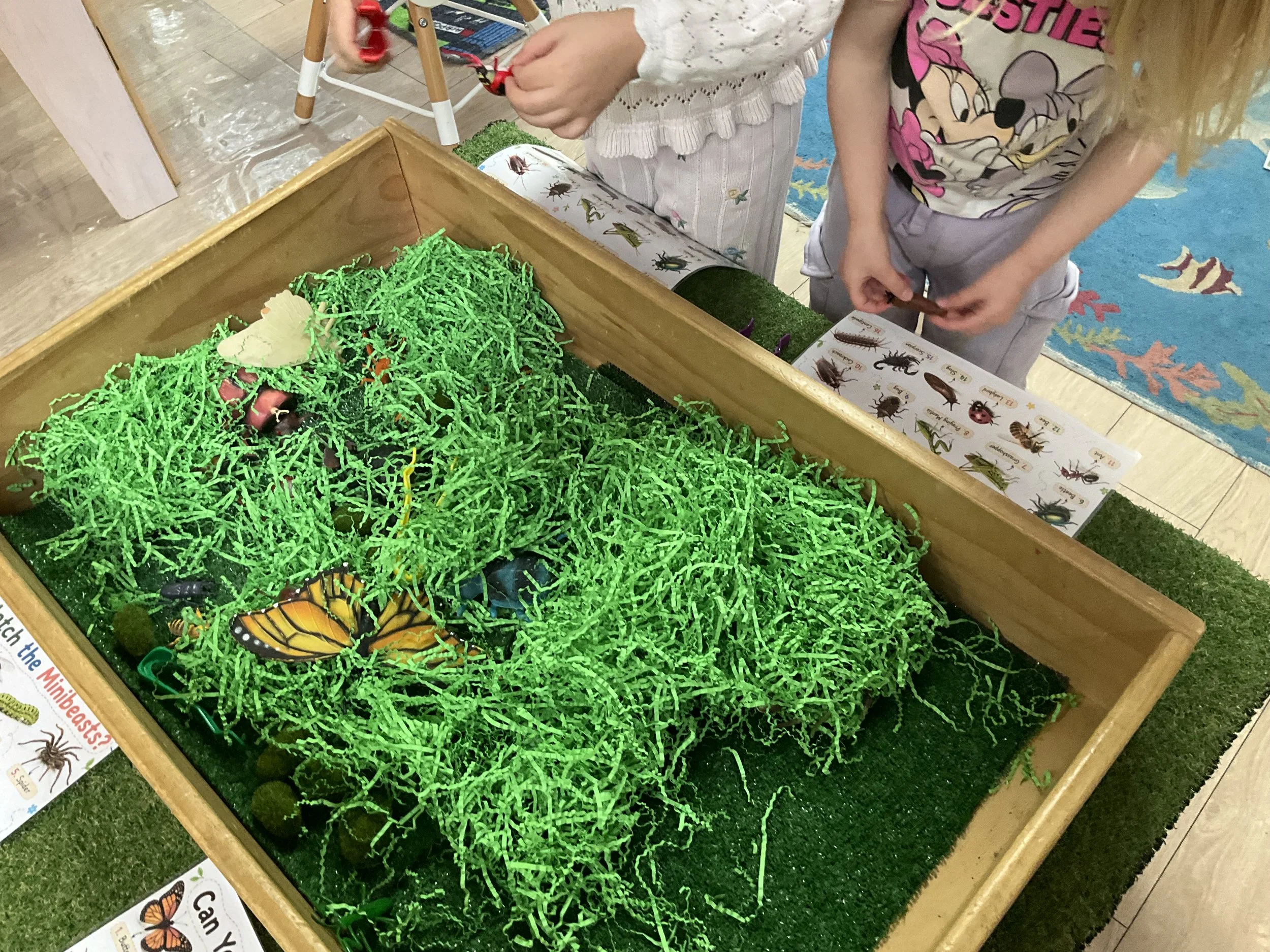 Children at a display table of plastic insects and bugs, with some green shredded paper simulating grass, and an insect identification chart nearby.