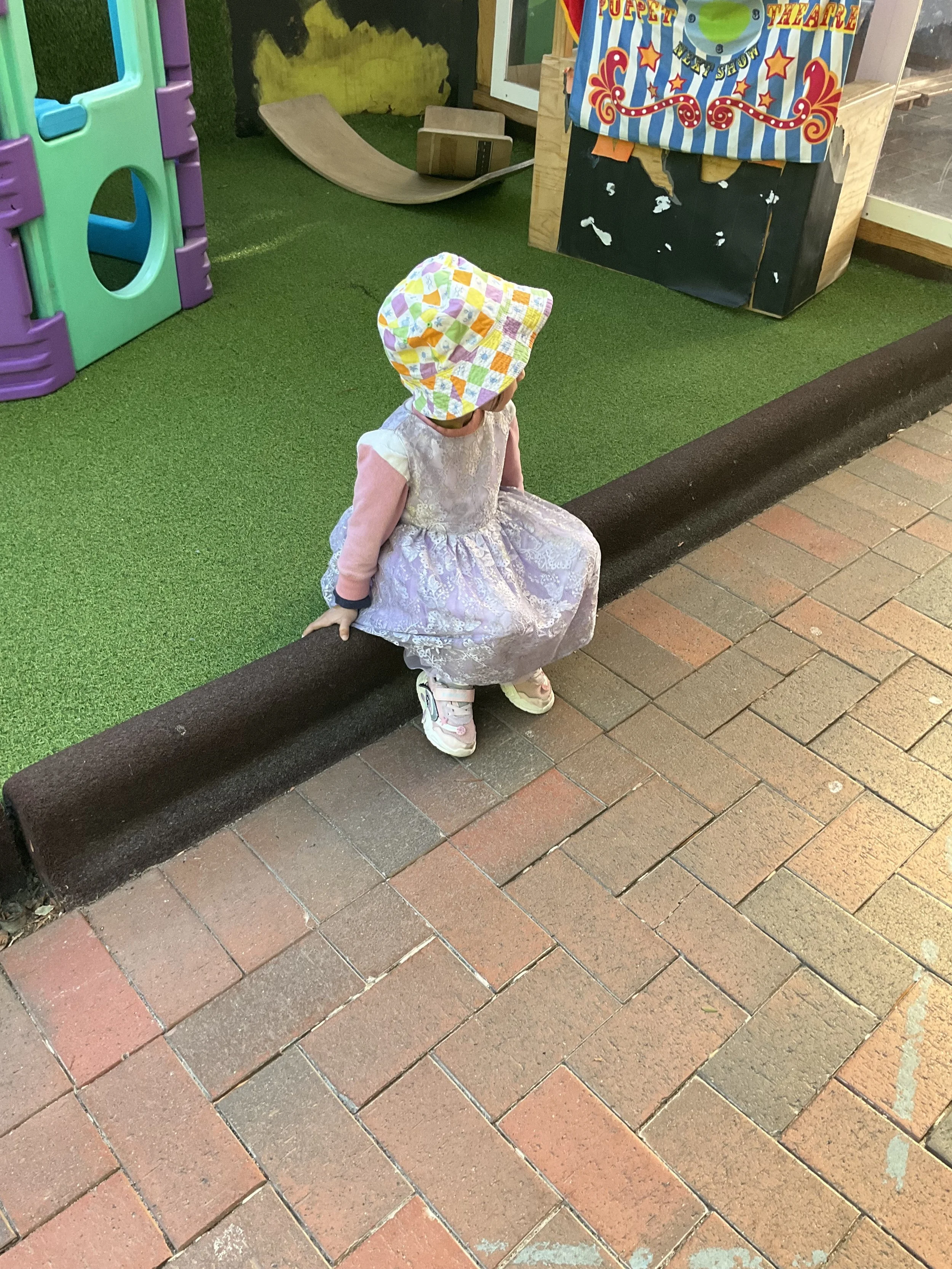 A young girl wearing a colorful checkered sun hat, a pink long-sleeve shirt, a lace dress, and sneakers sits on a brown curb outside a play area with green artificial turf. 